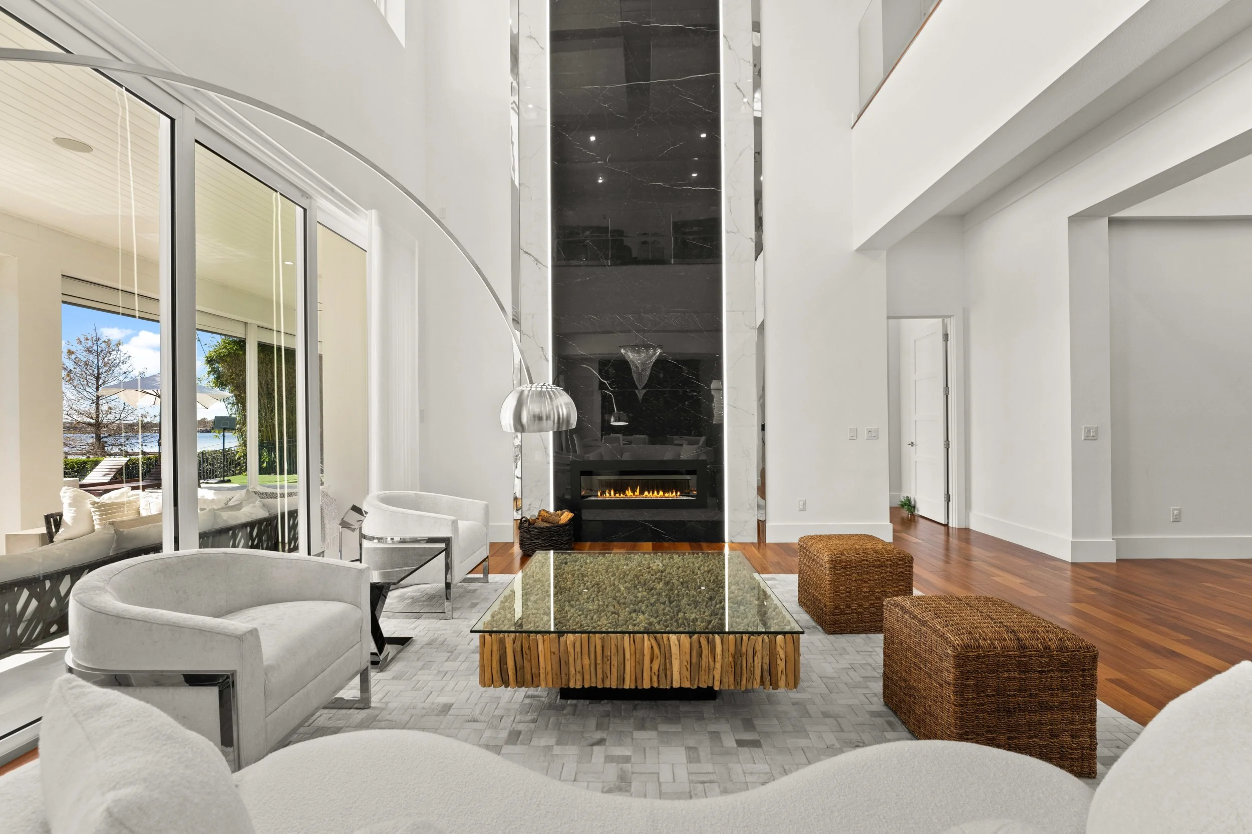 Modern living room with white walls, a large black marble fireplace, and a glass-top coffee table. There are white upholstered chairs, woven ottomans, and sliding glass doors leading to an outdoor area with outdoor furniture and a view of a lake or river.