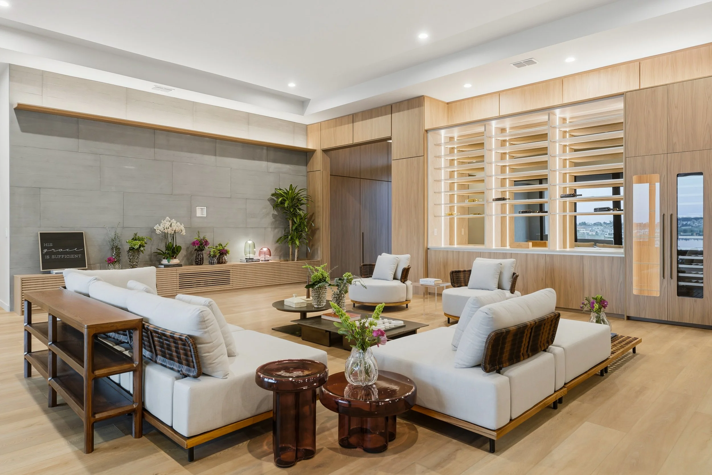 Modern living room with white sofas, wooden furniture, decorative plants, floral arrangements, and a gray stone wall with shelves.