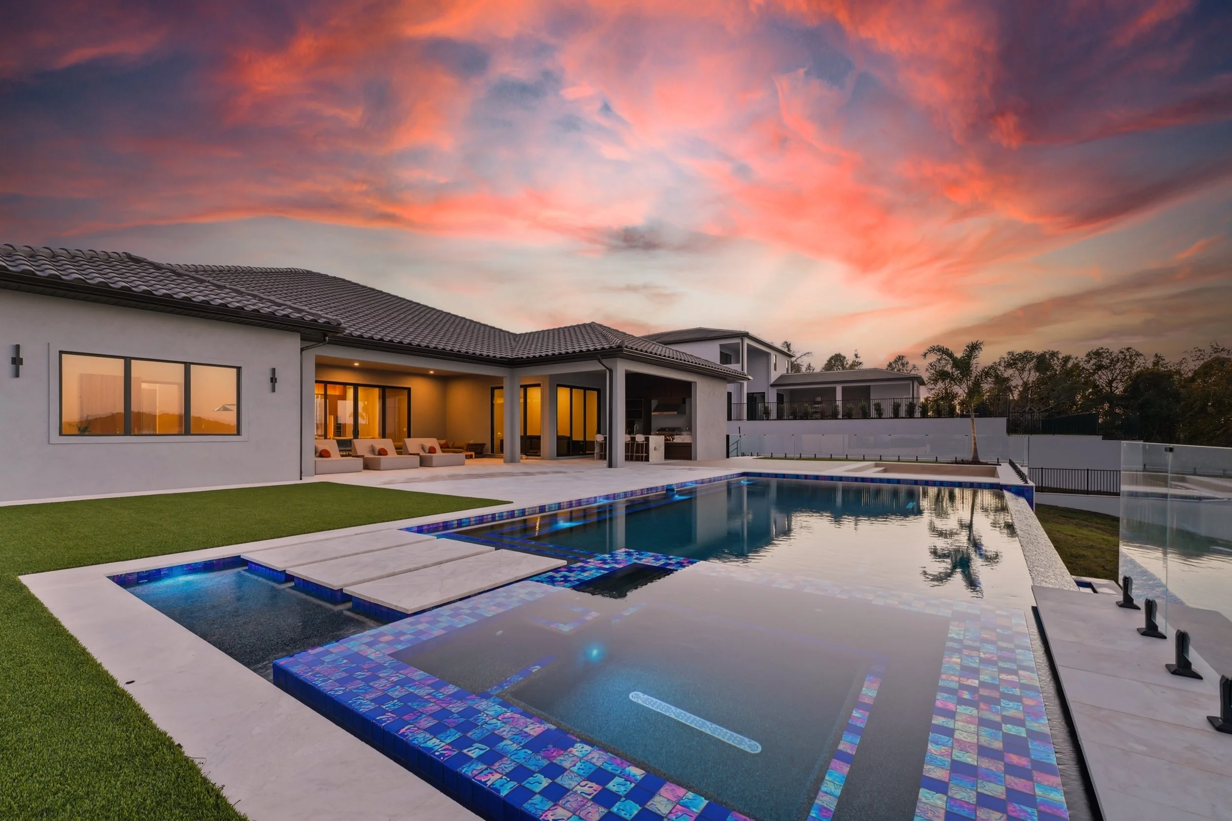 Modern backyard with a swimming pool and hot tub, patio with lounge chairs, and a house with large windows at sunset, colorful sky