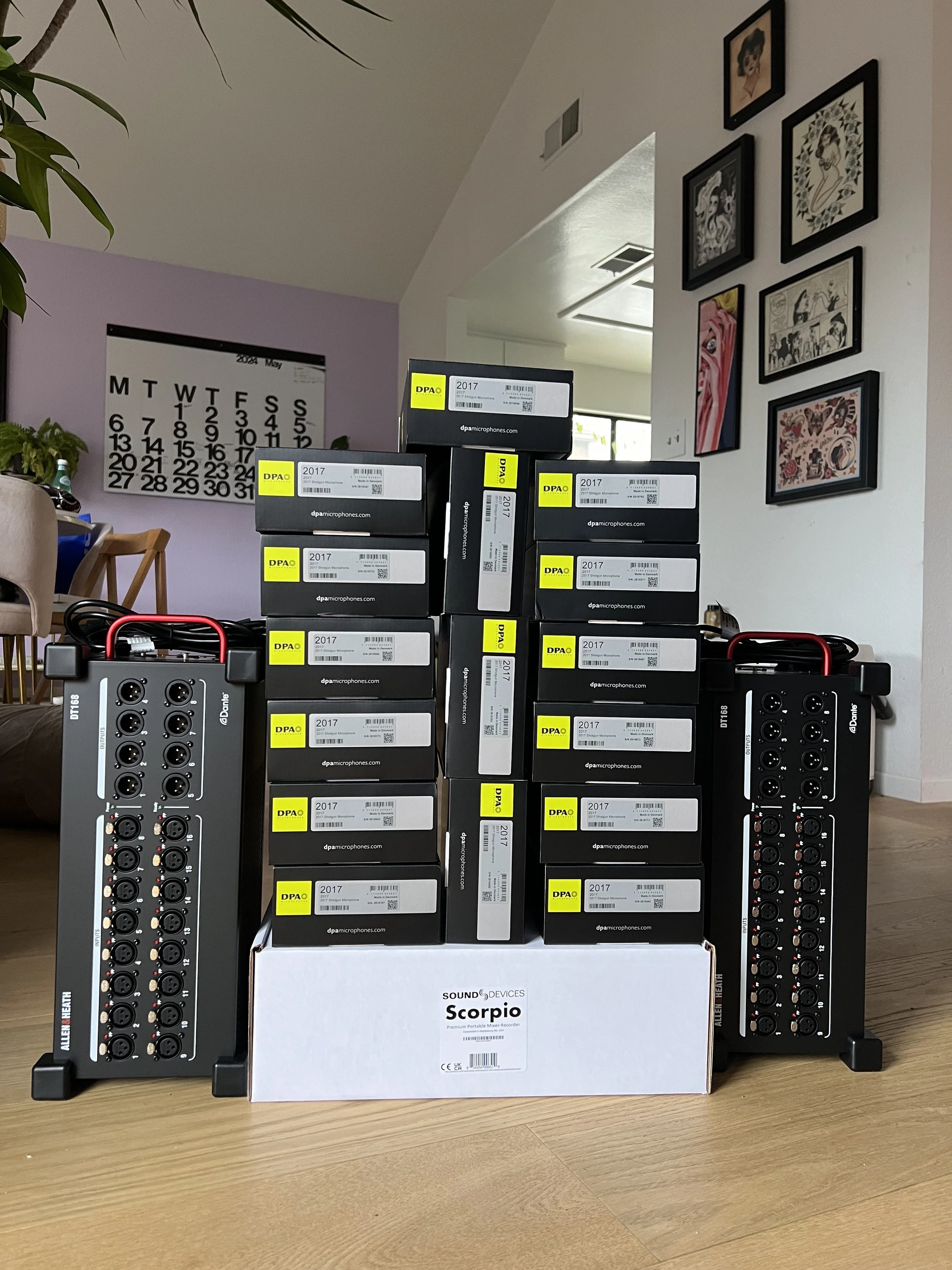 Multiple boxed DPA microphones stacked on top of a Sound Devices Scorpio portable recorder, with two Allen & Heath mixer units at either side, on a wooden table in a living room with wall art and a calendar in the background.