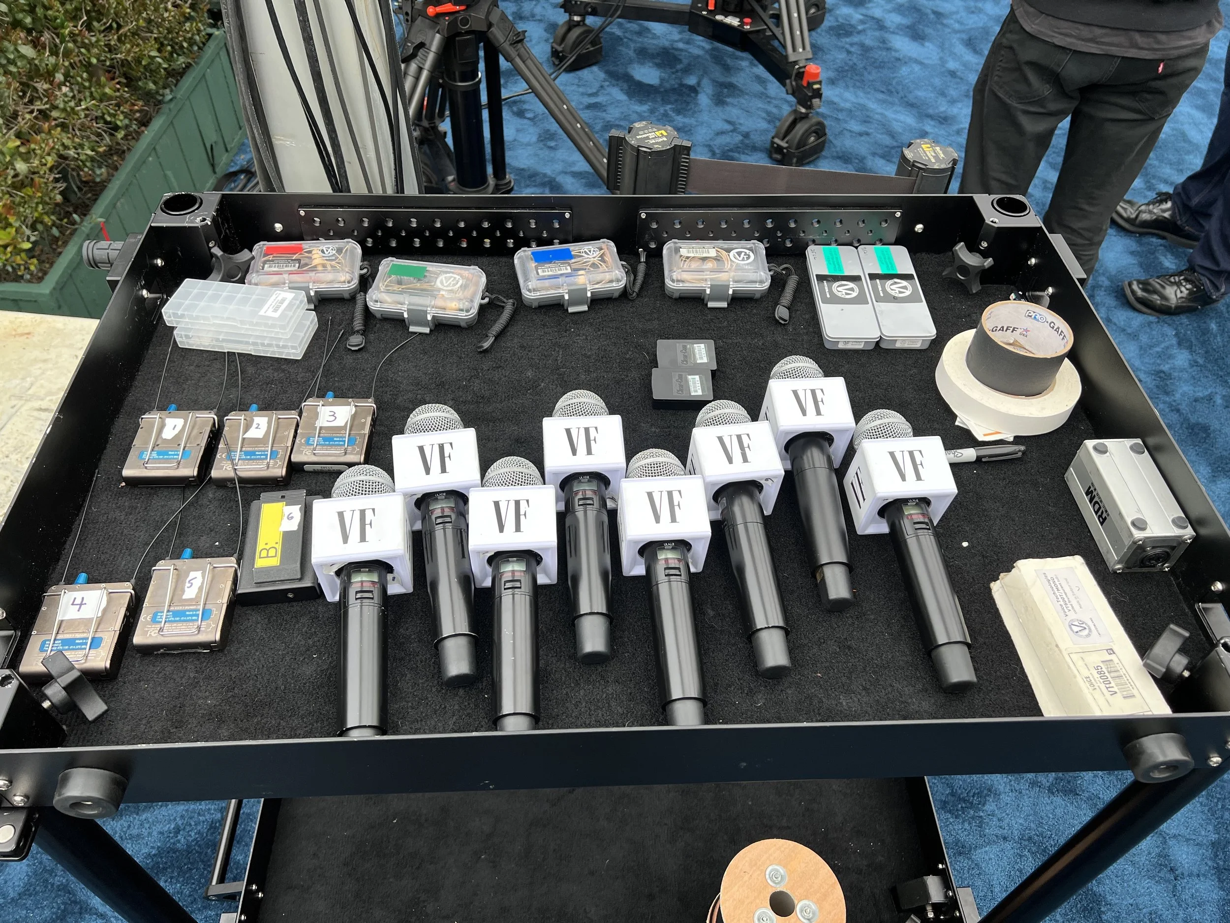 A table with multiple microphones labeled Vanity Fair, several small boxes with wires, and various equipment, with a blue carpet and some celebrities standing nearby.