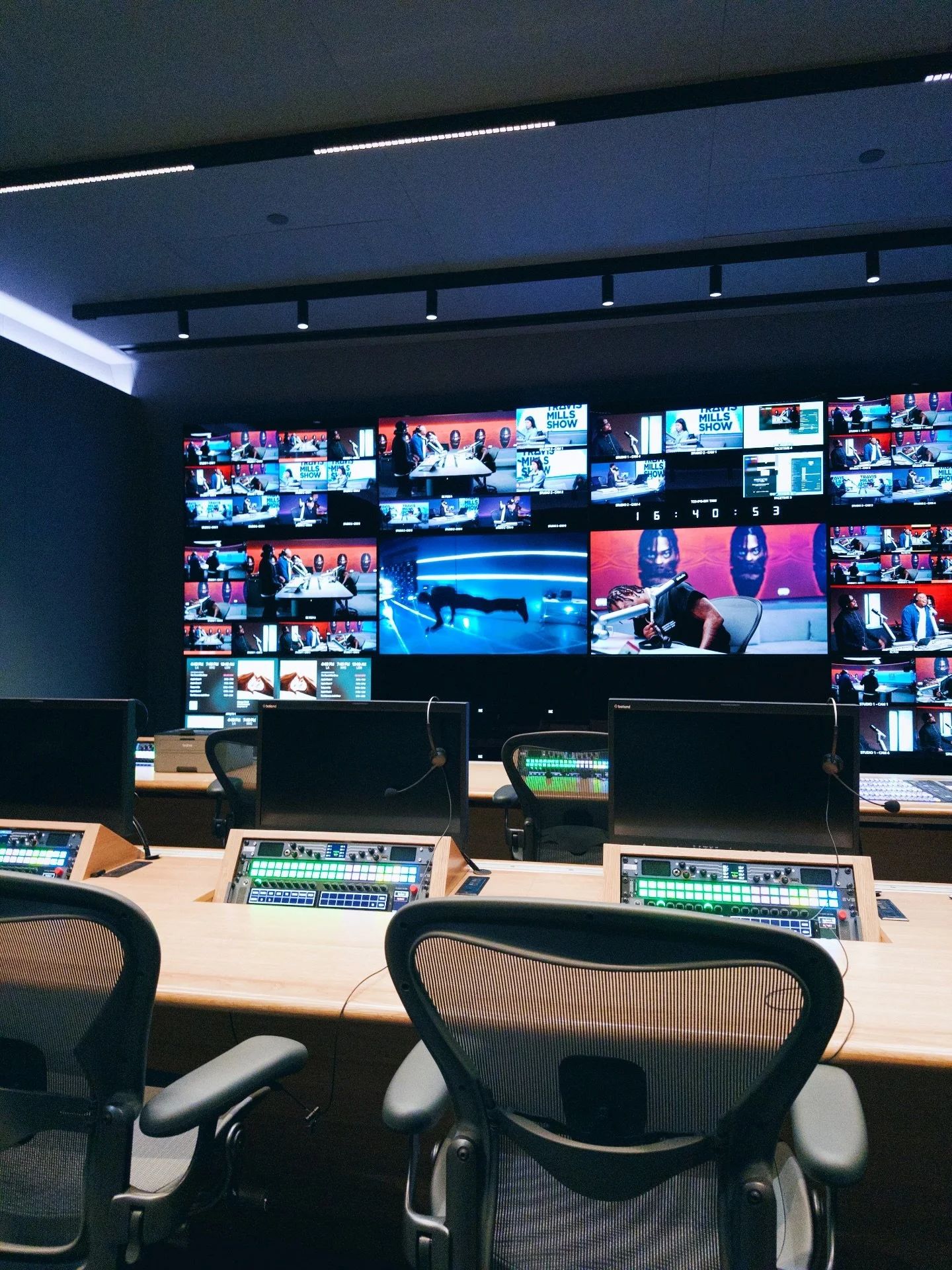 A control room with multiple computer screens and chairs, displaying a large monitor wall with various video feeds and a production setup.
