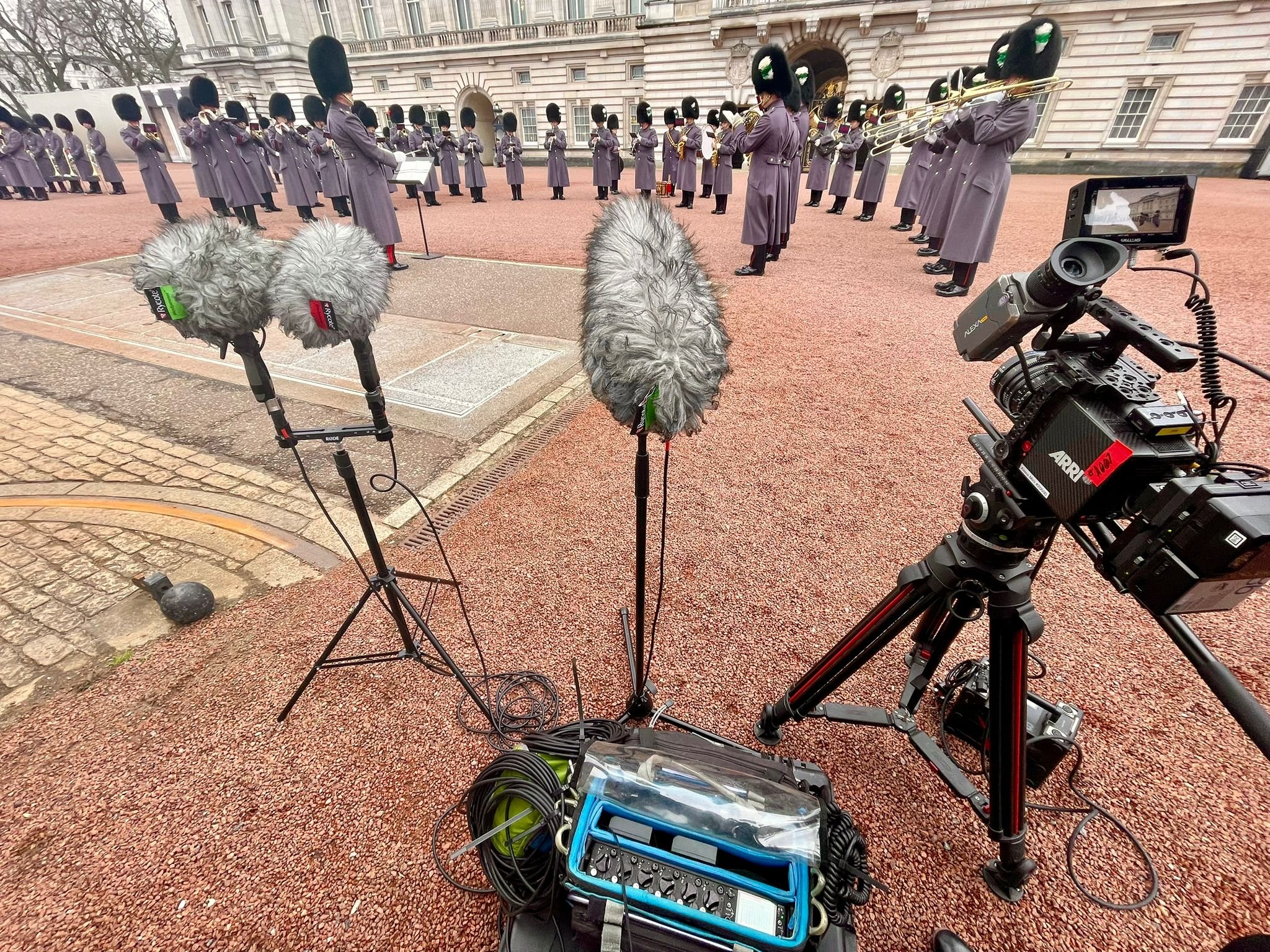 Camera equipment and microphones set up in front of Buckingham Palace, with the changing of the guards, officers dressed in purple uniforms and tall black hats playing instruments in front of a historic building.