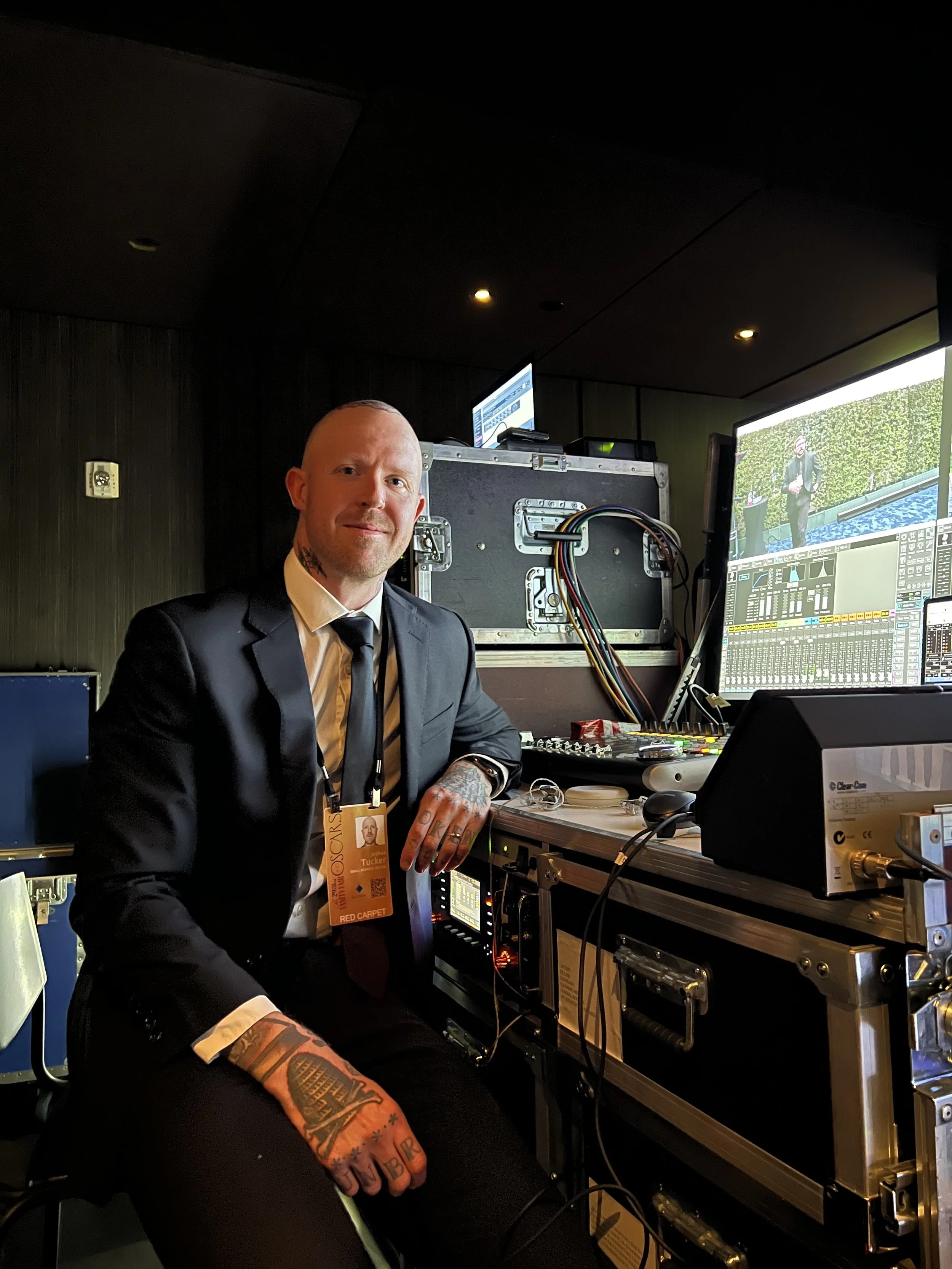 A man in a black suit and tie sitting at a video or TV control station with multiple monitors and equipment, wearing an event badge, tattoos visible on his neck and on his hands.