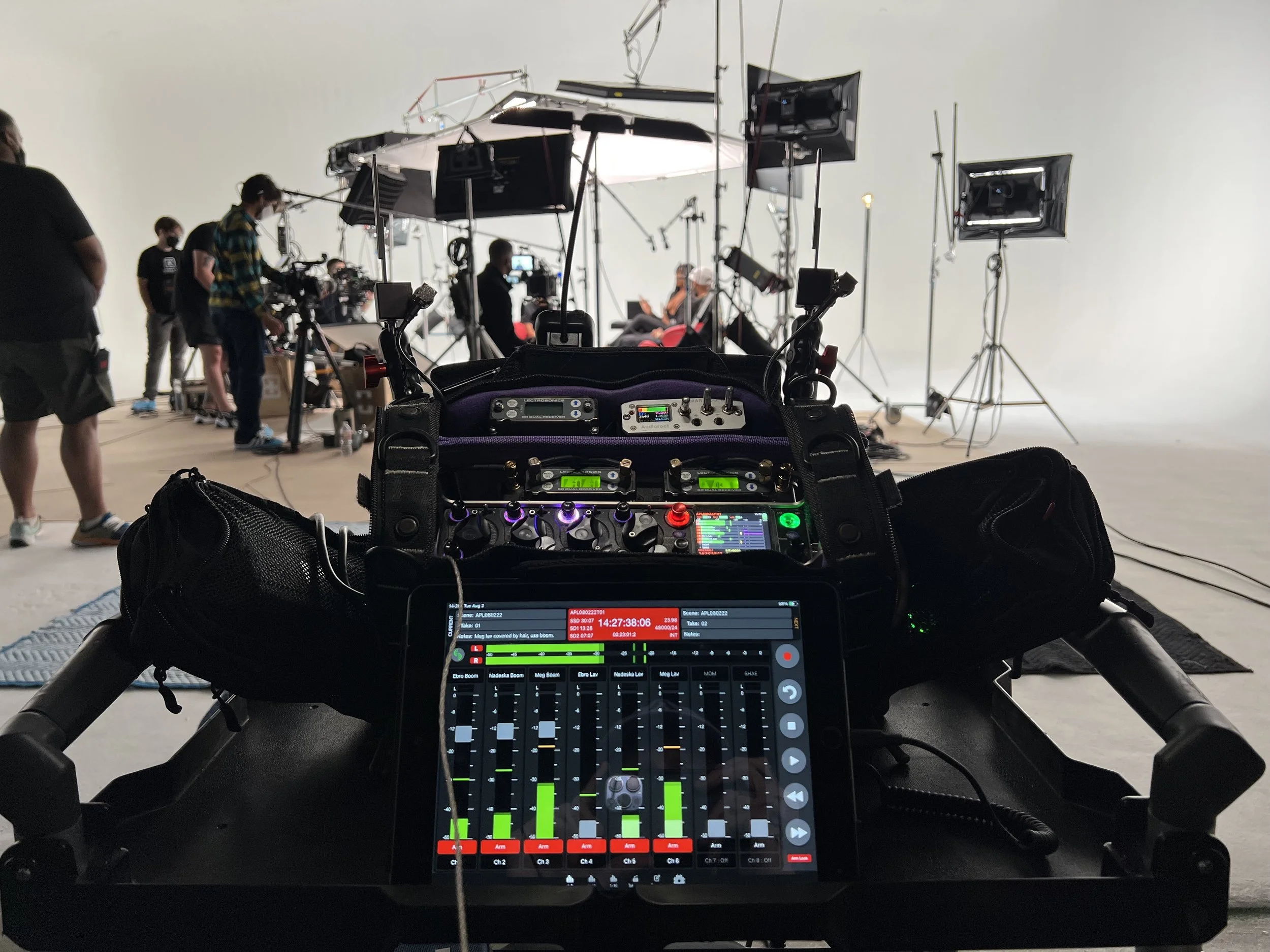 Close-up view of a digital audio mixing console with a touchscreen display showing audio levels, set on a black cart, with a professional video camera and audio equipment in a film or television studio behind it, crews setting up for filming.