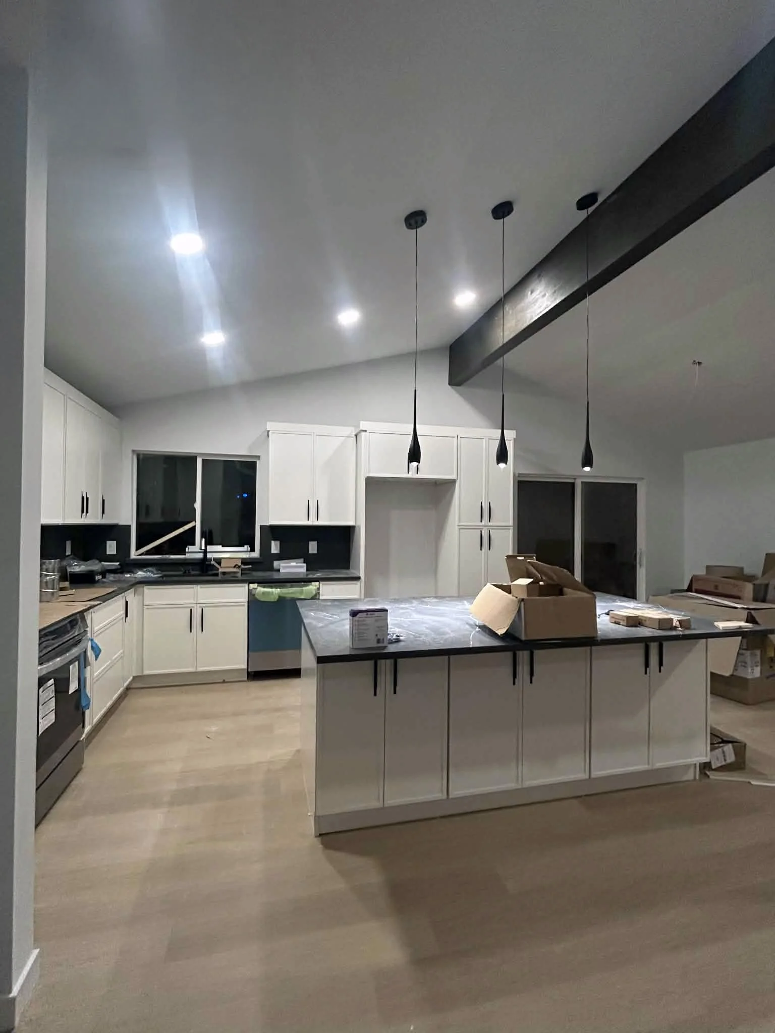 A modern kitchen under construction with white cabinets, a kitchen island, black pendant lights, and some boxes and tools on the island.