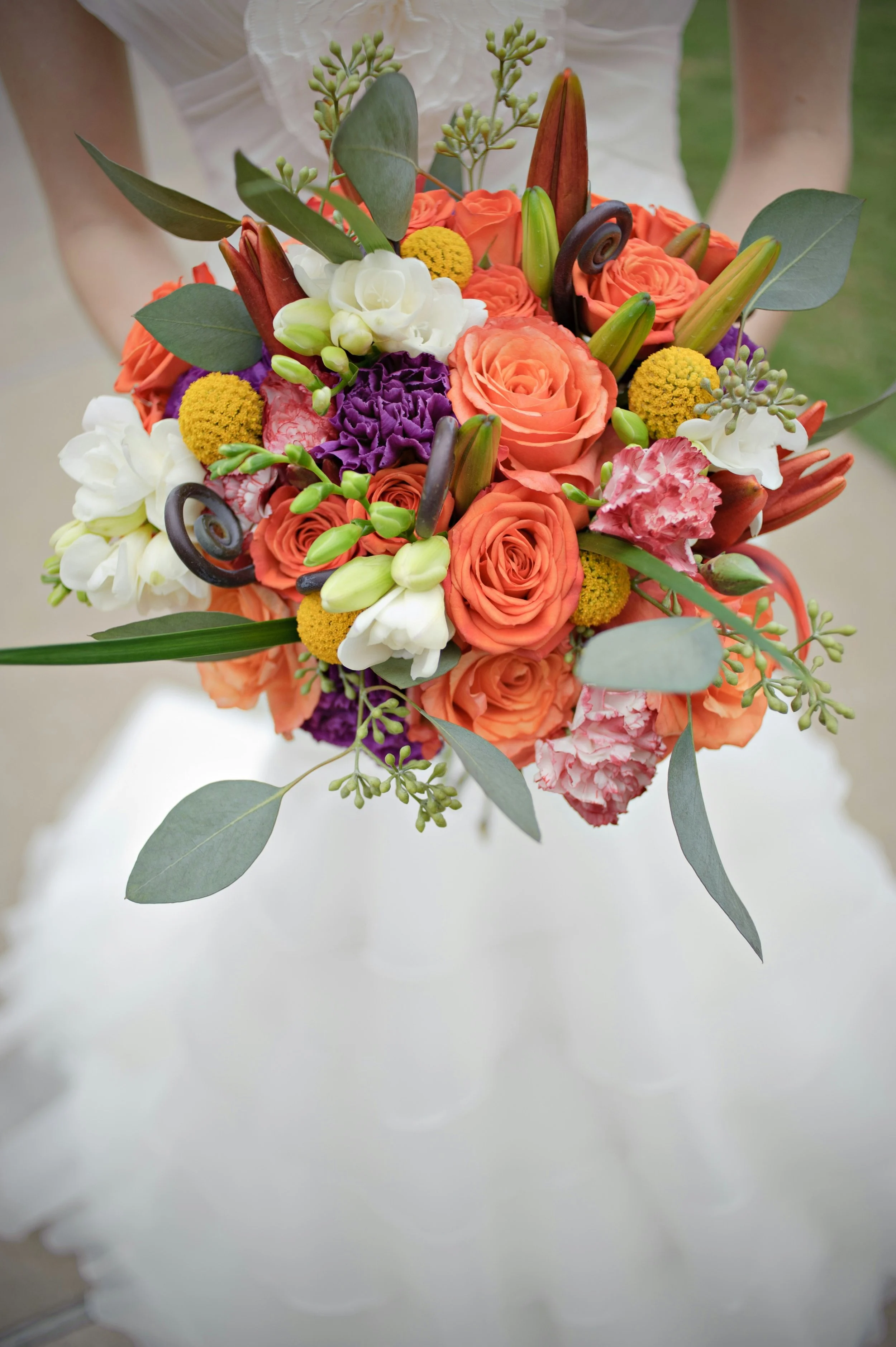 A colorful bouquet of flowers including roses, peonies, lilies, and greenery, held by a person in a white dress.