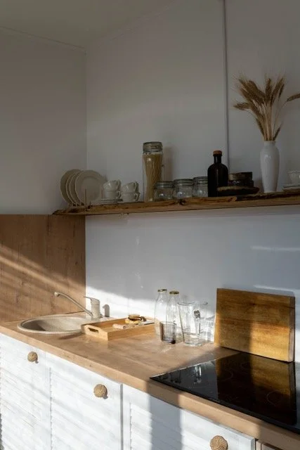 Kitchen countertop with glass jars, drinking glasses, a wooden cutting board, and a sink. Shelves above hold dishes, cups, a glass jar, a bottle, and a white vase with dried plants.