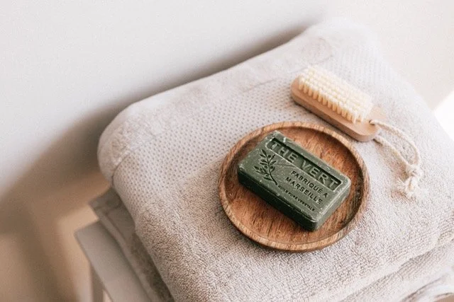 Wooden tray with a green bar of soap labeled 'The Vert Fabrique à Marseille' and a light-colored soap brush, placed on a folded white towel.