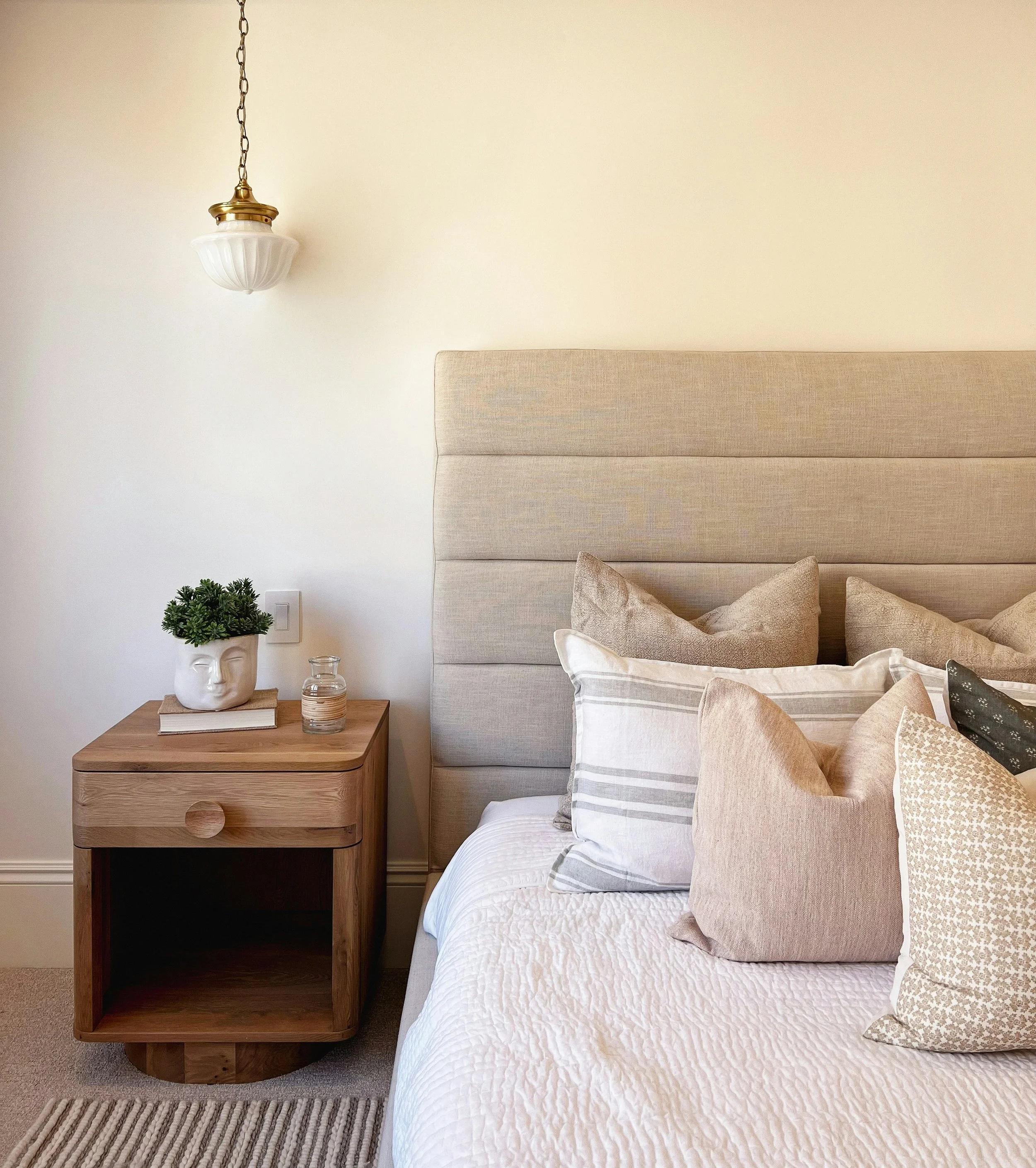 A beige upholstered bed with multiple pillows, a wooden nightstand with a potted plant, a book, two glasses, and a striped rug in front of the bed, against a plain off-white wall with a hanging vintage-style white and gold pendant light.