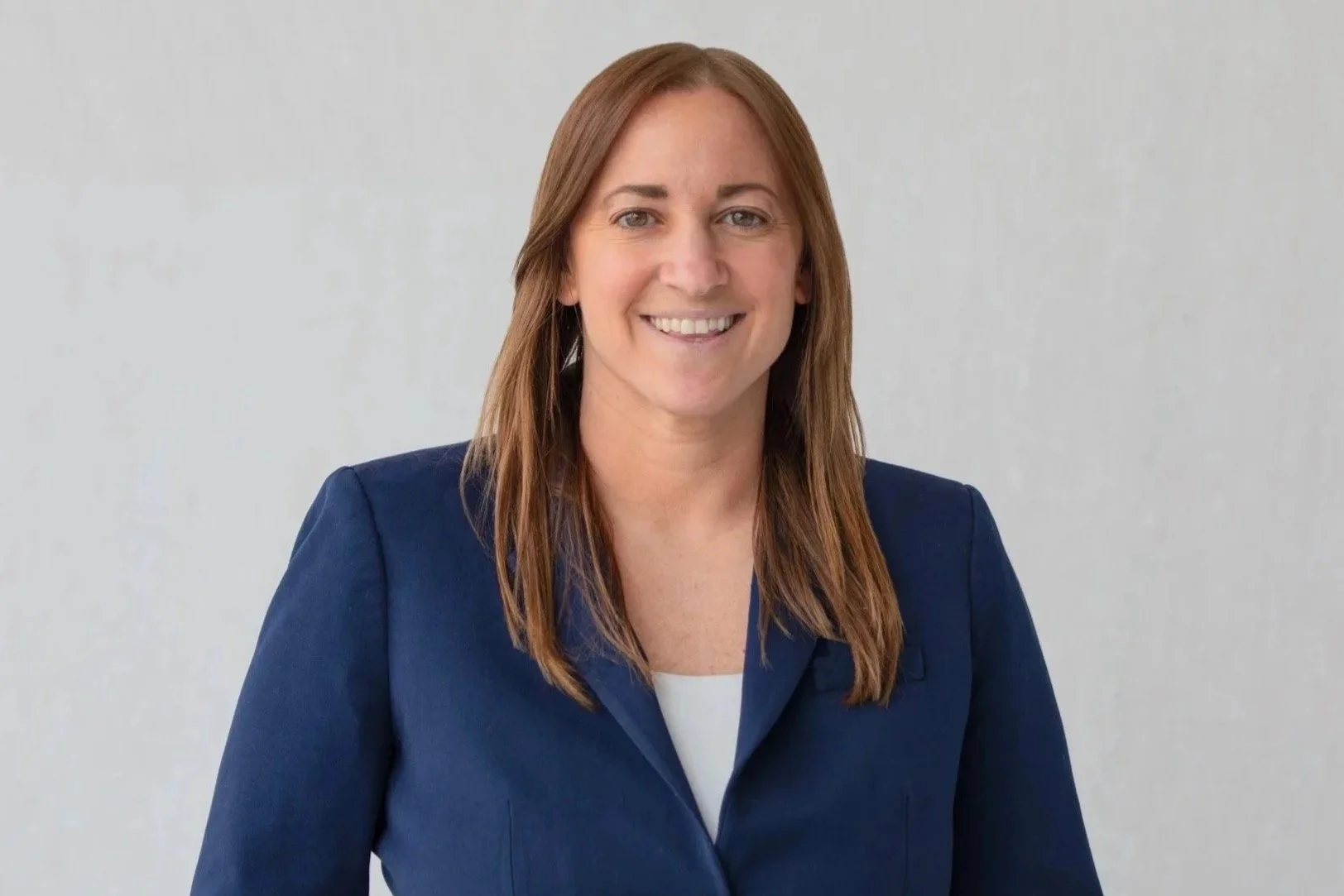 A woman with shoulder-length brown hair, wearing a navy blazer and a white top, smiling against a plain light gray background.