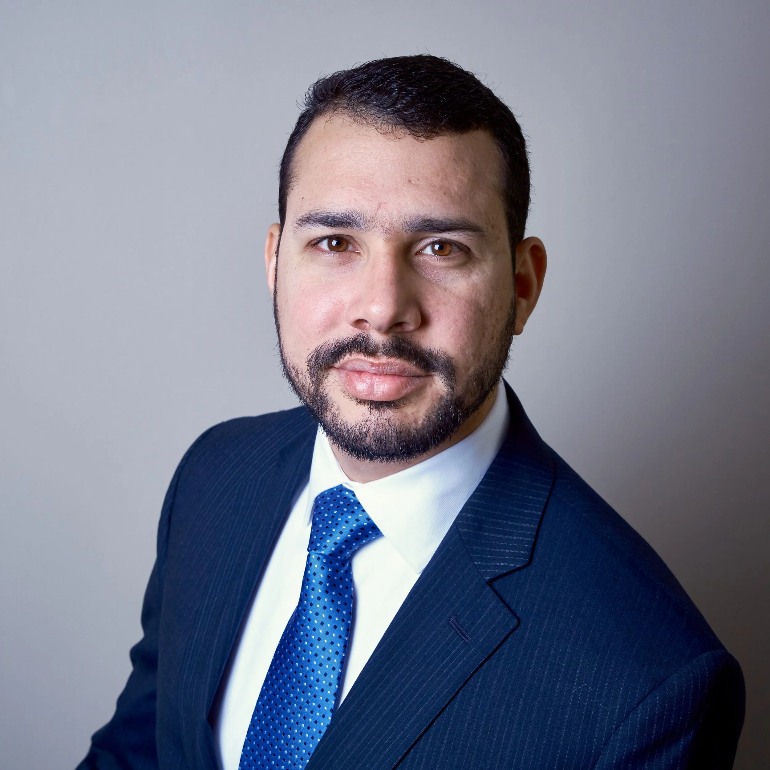 Headshot of a man with dark hair, beard, wearing a dark suit, white shirt, and blue tie, against a plain background.