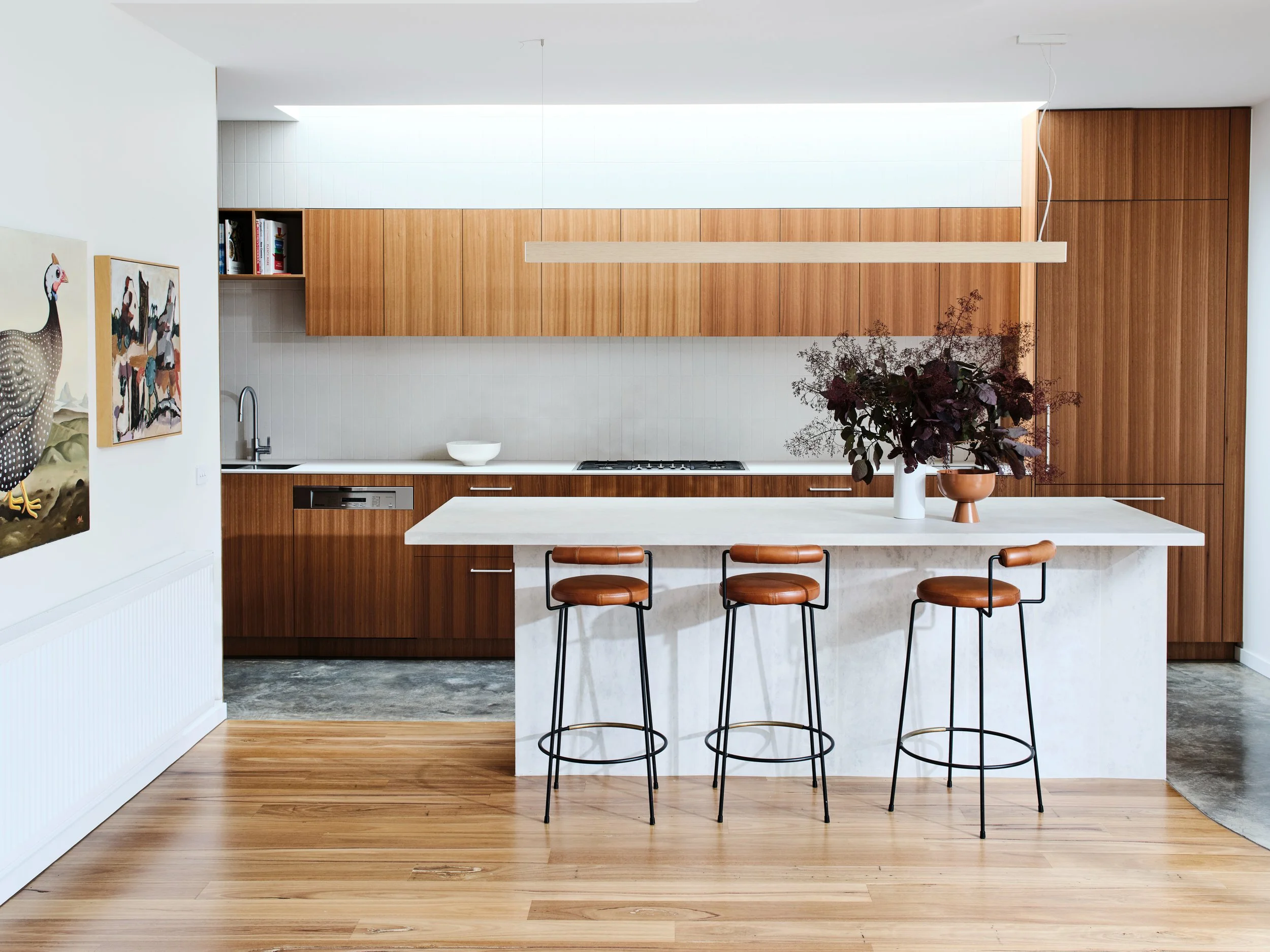 Modern kitchen with white tiled splashback, wooden cabinets, white marble island with three leather bar stools, artwork on the wall, and a large plant in a white vase.