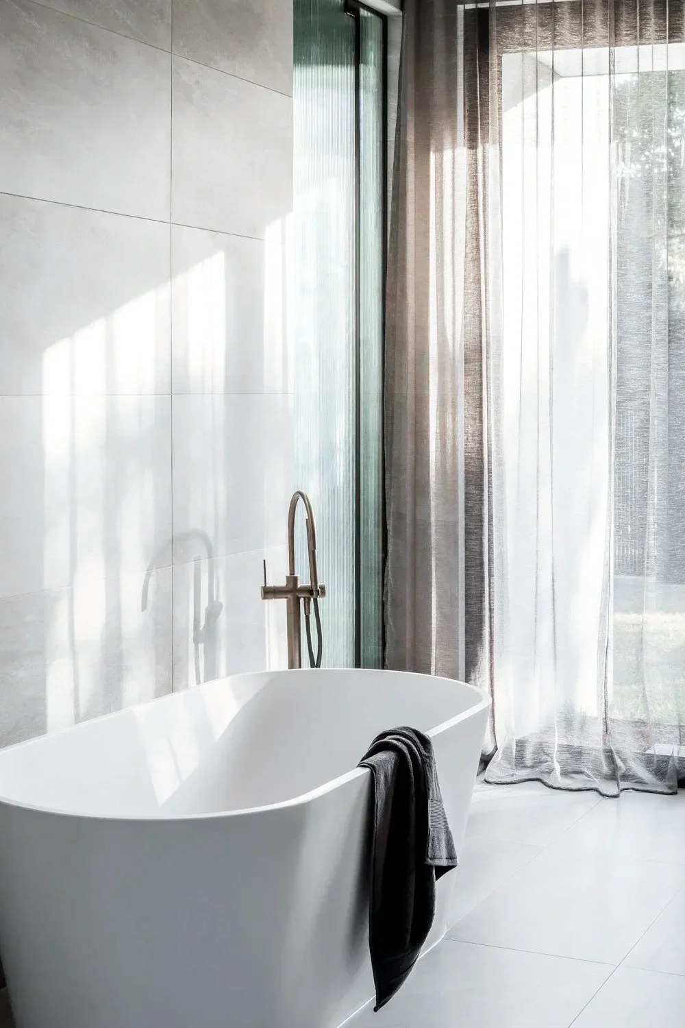 A modern tiled bathroom with a white freestanding bathtub near large windows with sheer curtains and a view of greenery outside. A towel hangs over the edge of the tub, and sunlight streams in, creating shadows on the light-colored tiled walls.