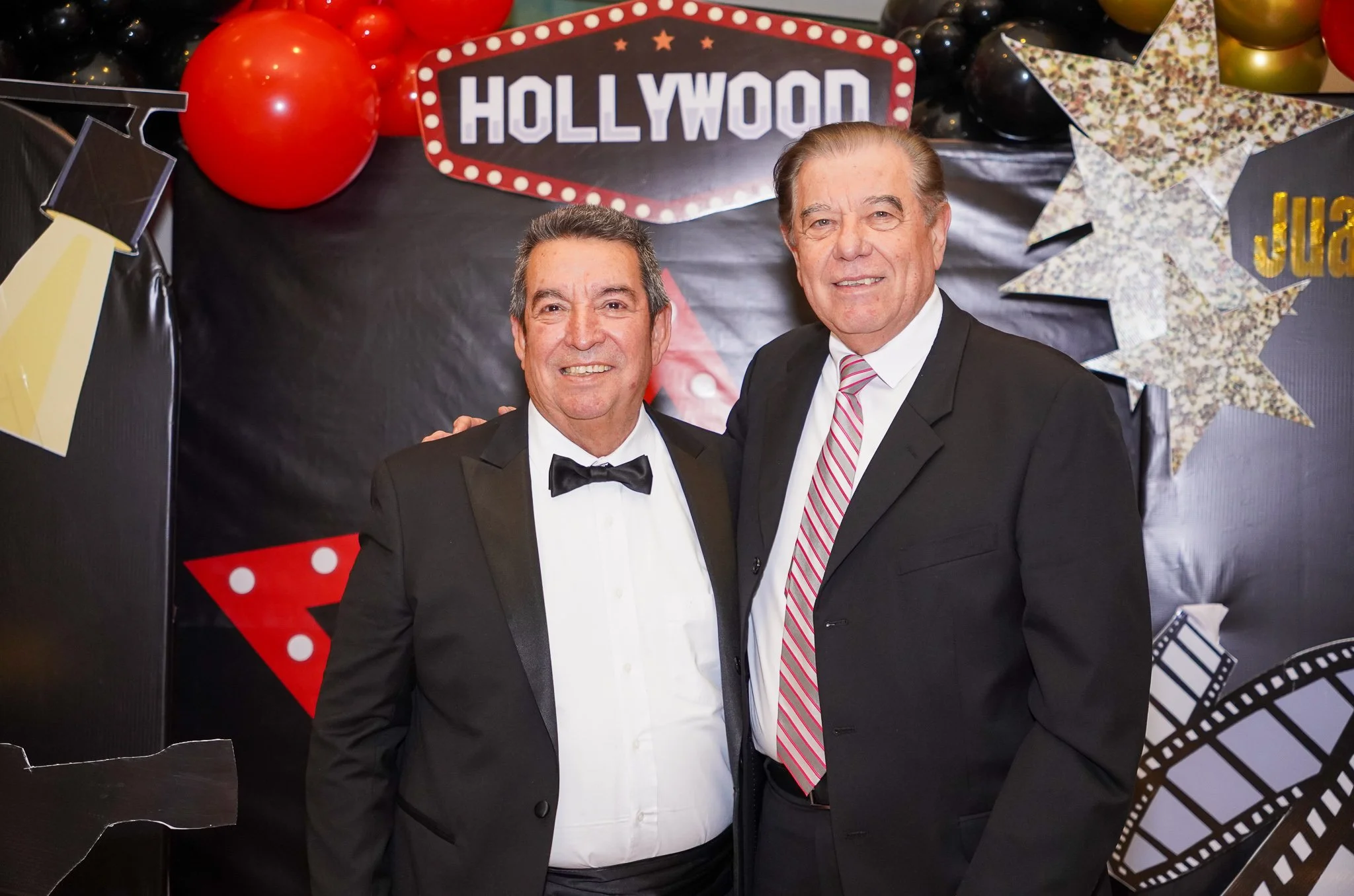 Dos hombres con trajes elegantes en una celebración con decoraciones de Hollywood, globos rojos, estrellas decorativas y un cartel que dice 'Hollywood'.