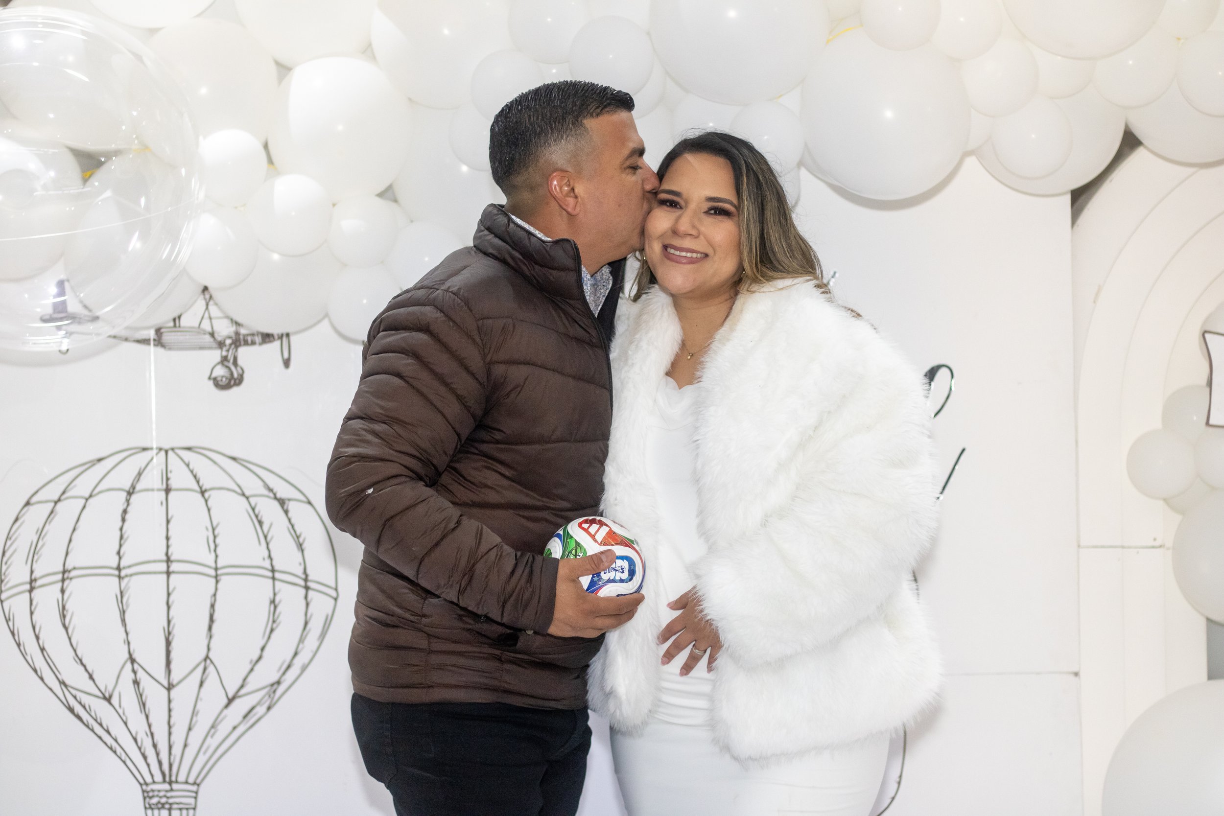 Una pareja en una celebración navideña, la mujer viste un abrigo blanco de peluche y el hombre lleva un suéter gris, ambos sosteniendo una pelota de fútbol con un fondo decorado con globos blancos y esquemas de globos en las paredes.