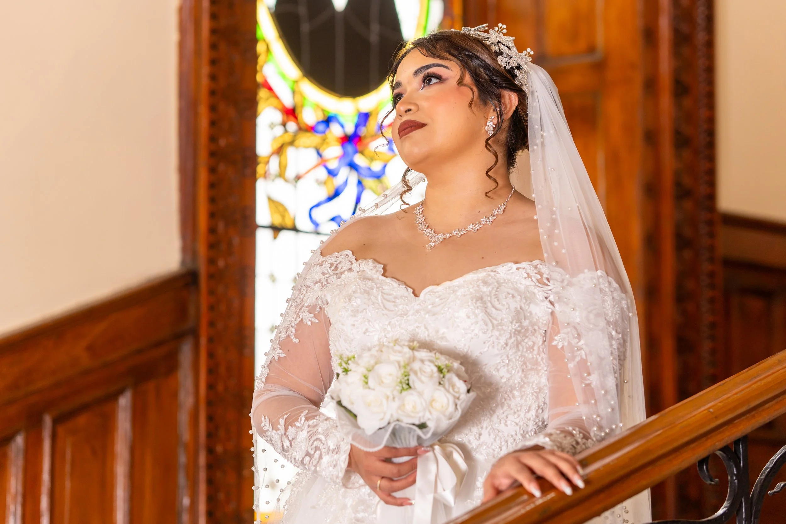 Mujer vestida de novia con velo y joyería, sosteniendo un ramo de flores blancas, en una iglesia con vitrales de colores.