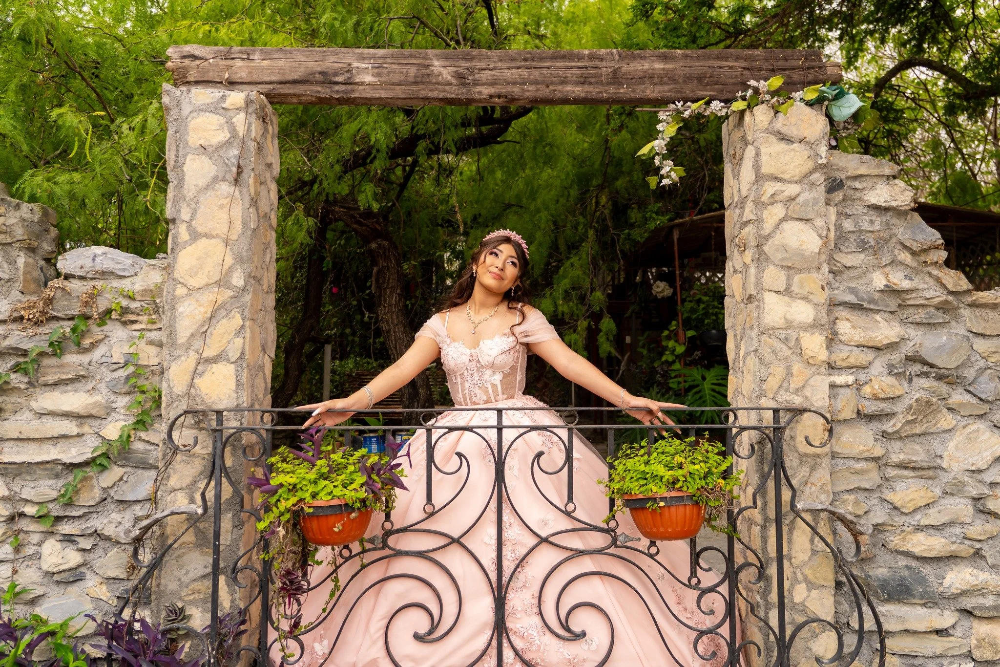 Joven mujer vestida con un vestido rosa y diadema, posa con las manos extendidas en una terraza de piedra con plantas colgantes, rodeada de árboles verdes.