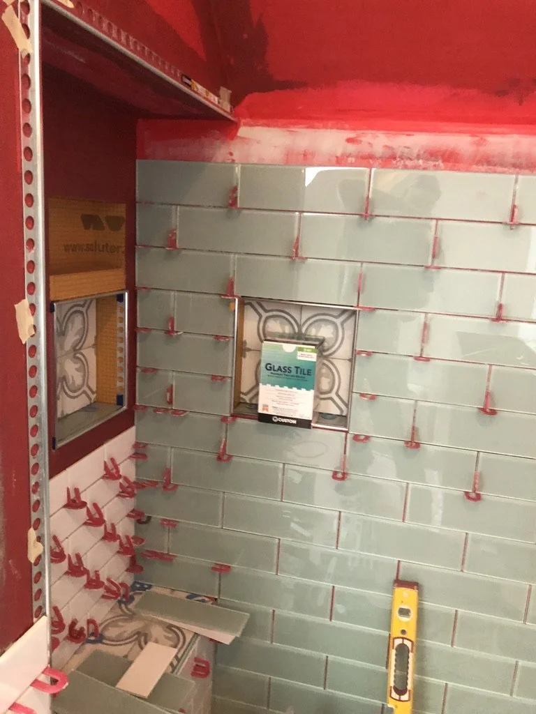 Interior of a store displaying a wall of light green glass tiles with red spacers, a small rectangular cutout in the tile wall, and a yellow level tool leaning against the wall.