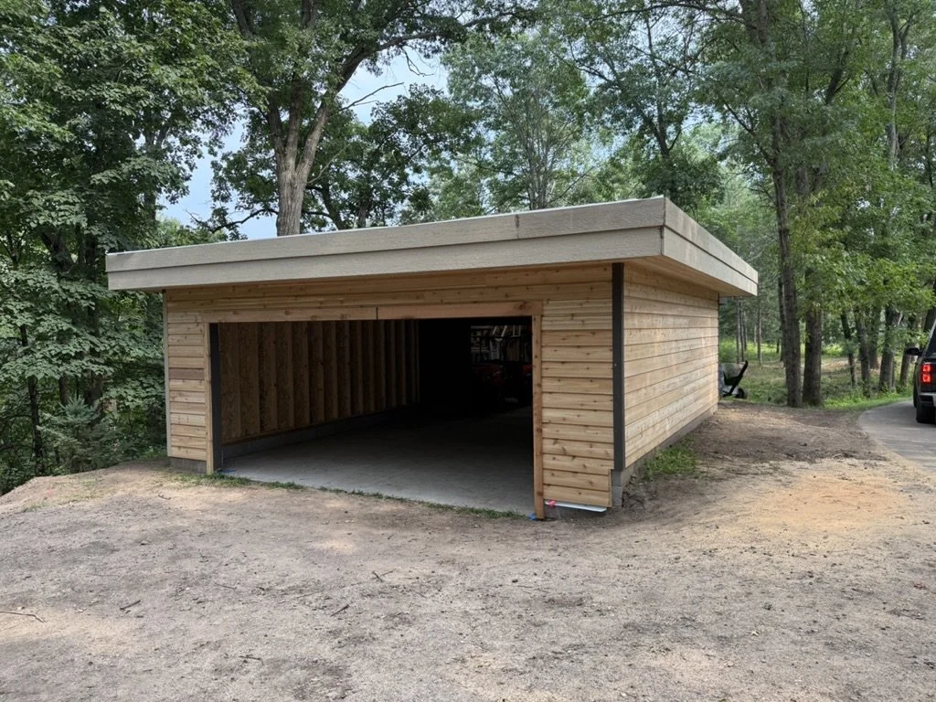 A wooden garage or carport with an open front, situated on a dirt and gravel driveway, surrounded by trees.