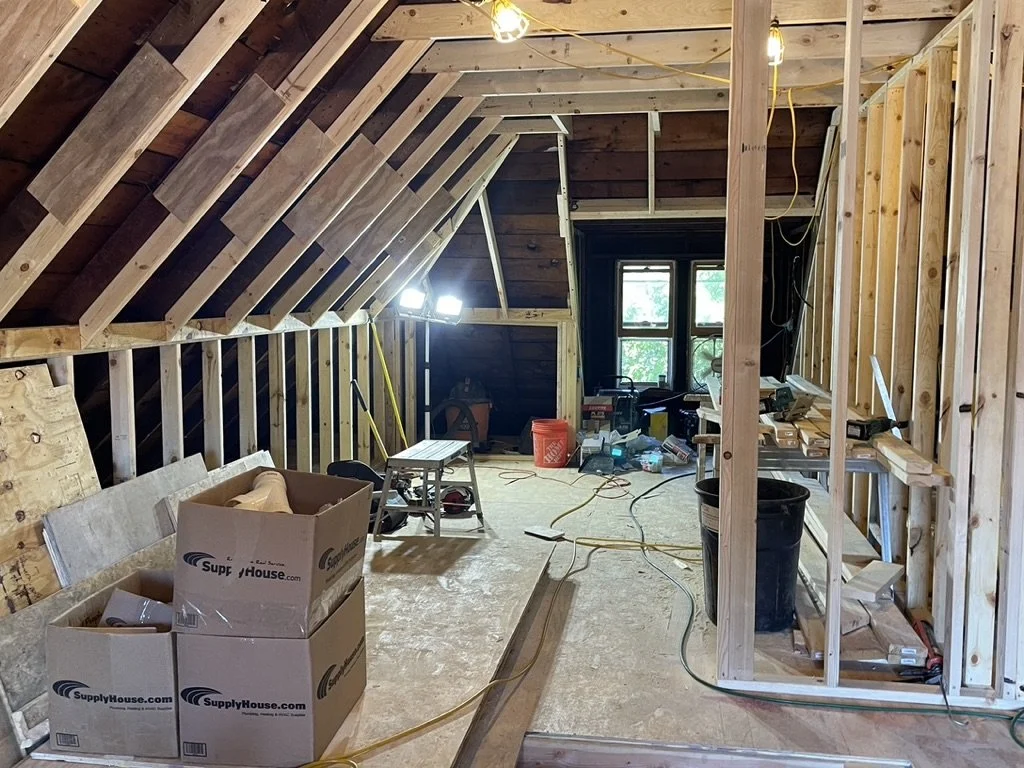 Attic under construction with unfinished wooden framing, some boxes, tools, and equipment scattered around, and a window at the far end.