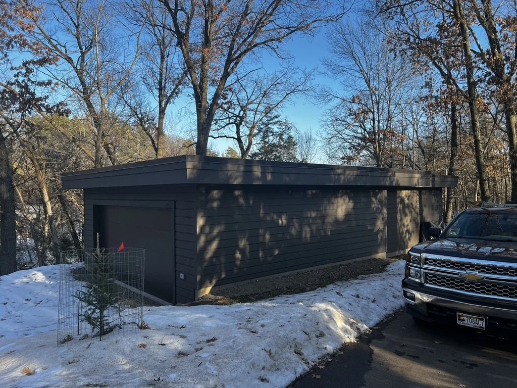 A black garage with a flat roof next to a parking lot, with a Chevrolet truck parked nearby. There are snow patches on the ground and trees in the background.