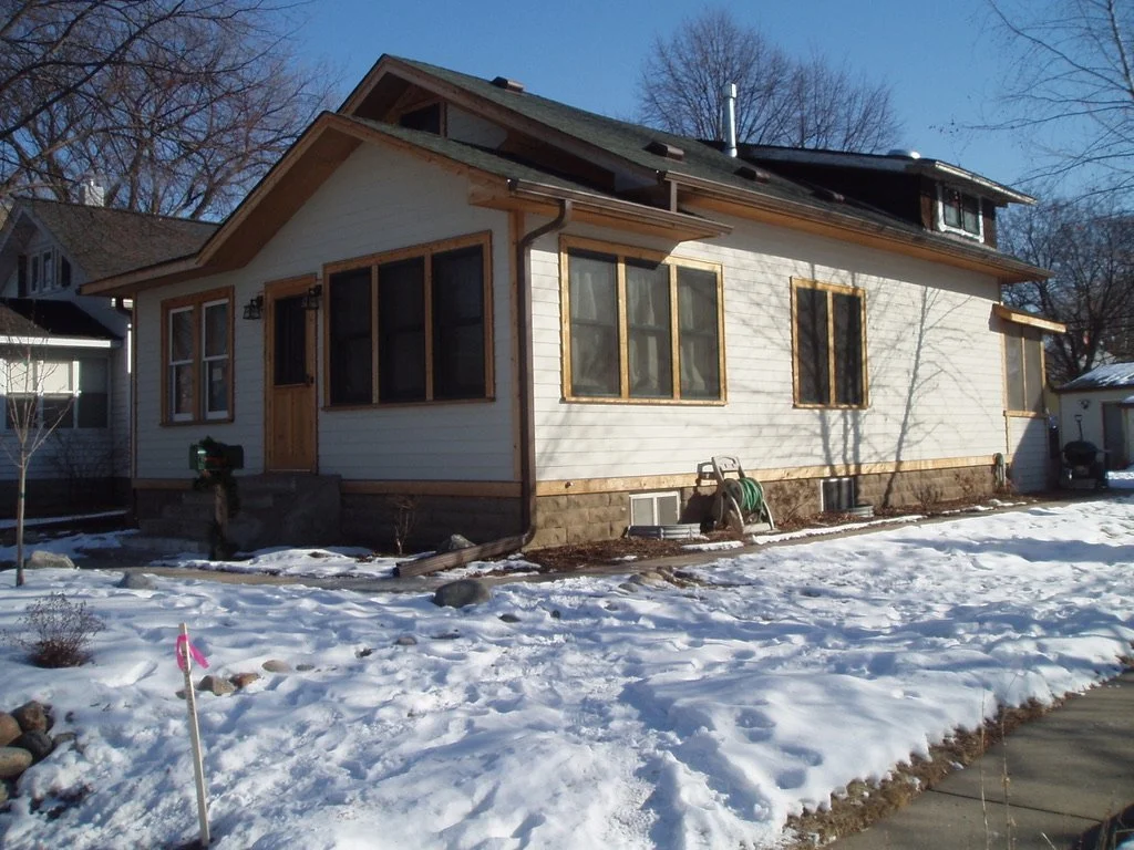 A house with white siding and a stone foundation, with new wooden window frames and a wooden front door, surrounded by snow in a neighborhood with leafless trees.