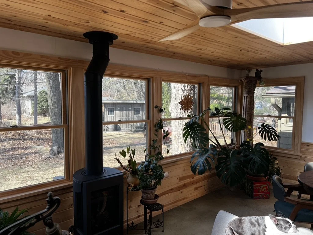 Living room with wood-paneled ceiling, multiple windows, plants, a black stove, and a glimpse of the outdoors.