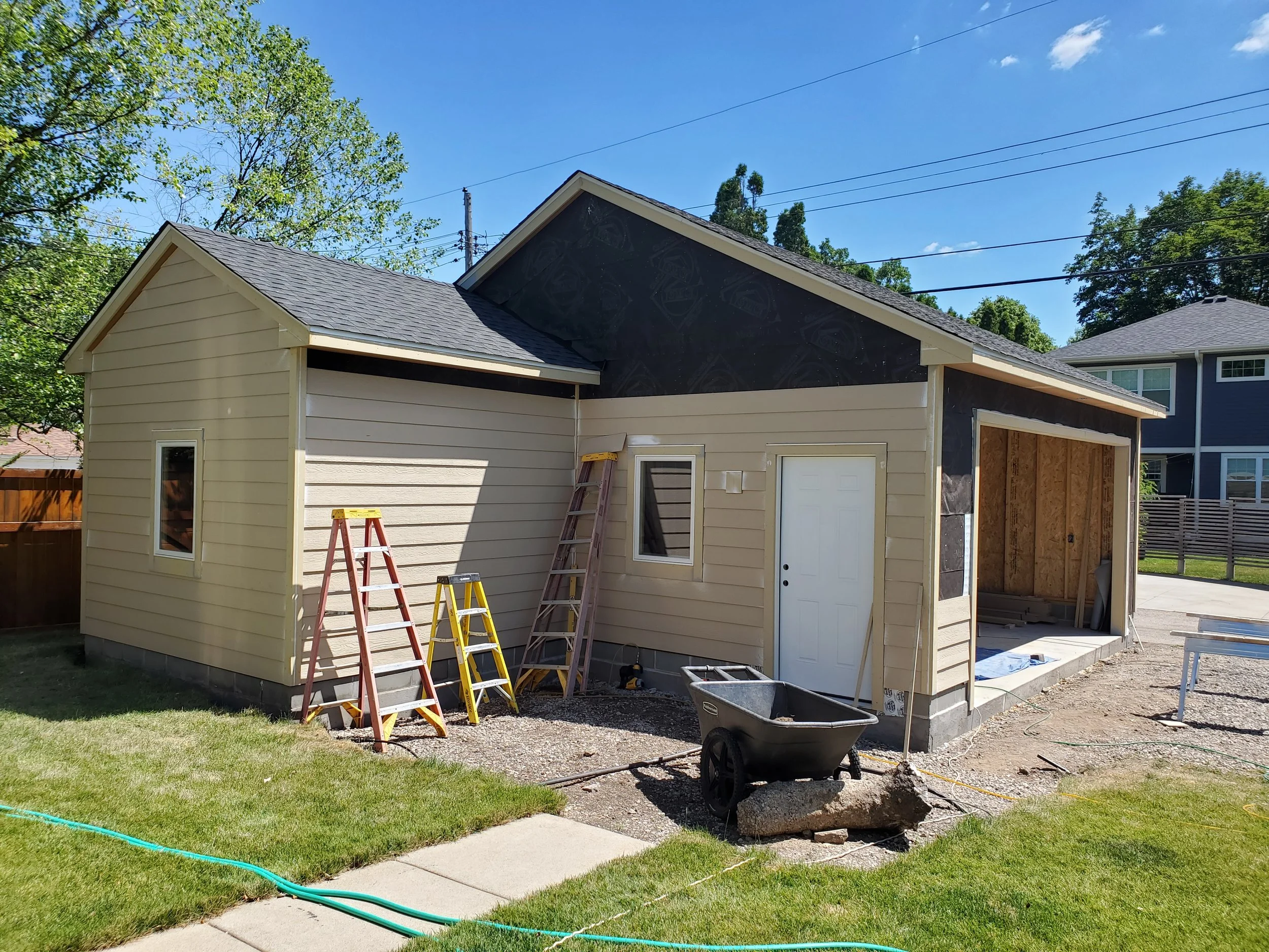 Side view of a house under construction with beige siding, windows, and an attached garage with partial wall framing. Ladders, a wheelbarrow, and building materials are present in the yard.
