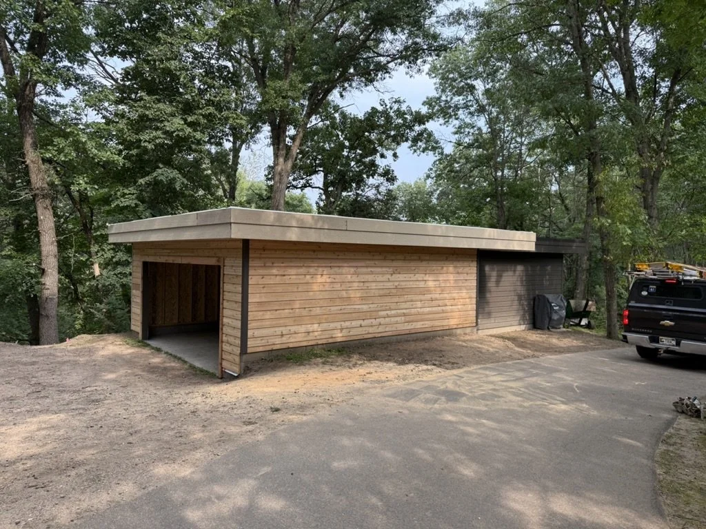 A small modern building with wooden exterior walls and a flat roof, located in a wooded area, with a driveway and a black pickup truck parked nearby.