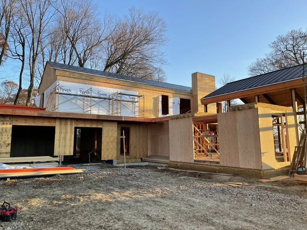 Construction site of a house with wooden framing and partially finished walls, trees in the background, and blue sky.