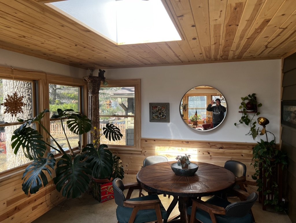 A cozy dining area with a round wooden table, surrounded by four chairs, within a room featuring wood-paneled walls and ceiling. Large windows offer a view of a backyard with trees. A round wall mirror and a small framed picture are on the wall. Ther