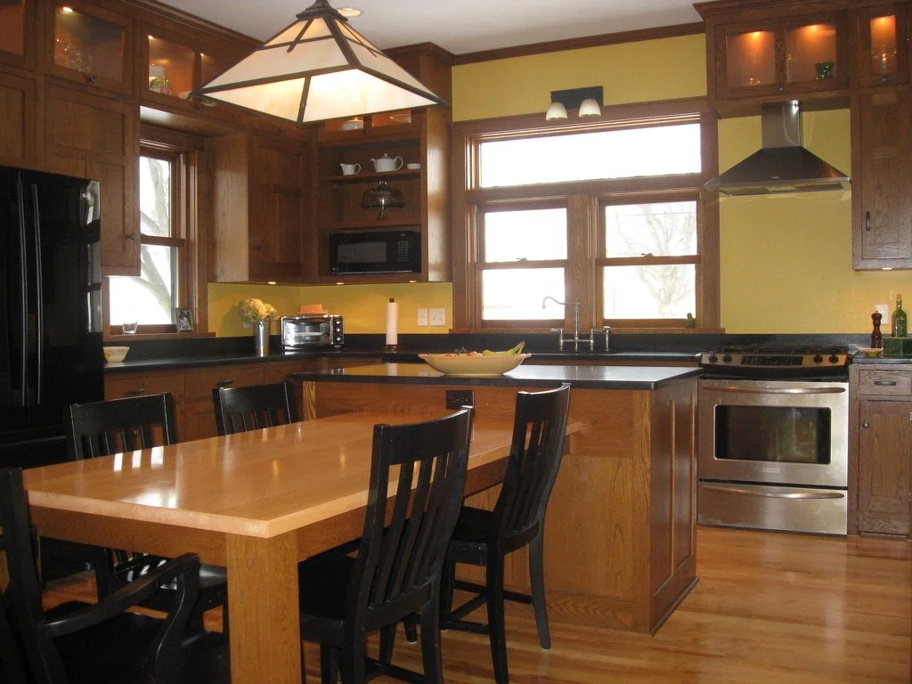 Kitchen with wooden cabinets, black countertops, stainless steel oven, microwave, black refrigerator, and a wooden dining table with black chairs.