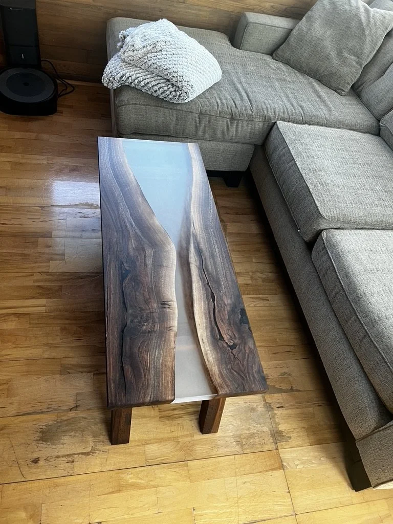 Living room with a gray sectional sofa, a folded blanket on the corner, and a wooden coffee table with a resin river design, on a hardwood floor.