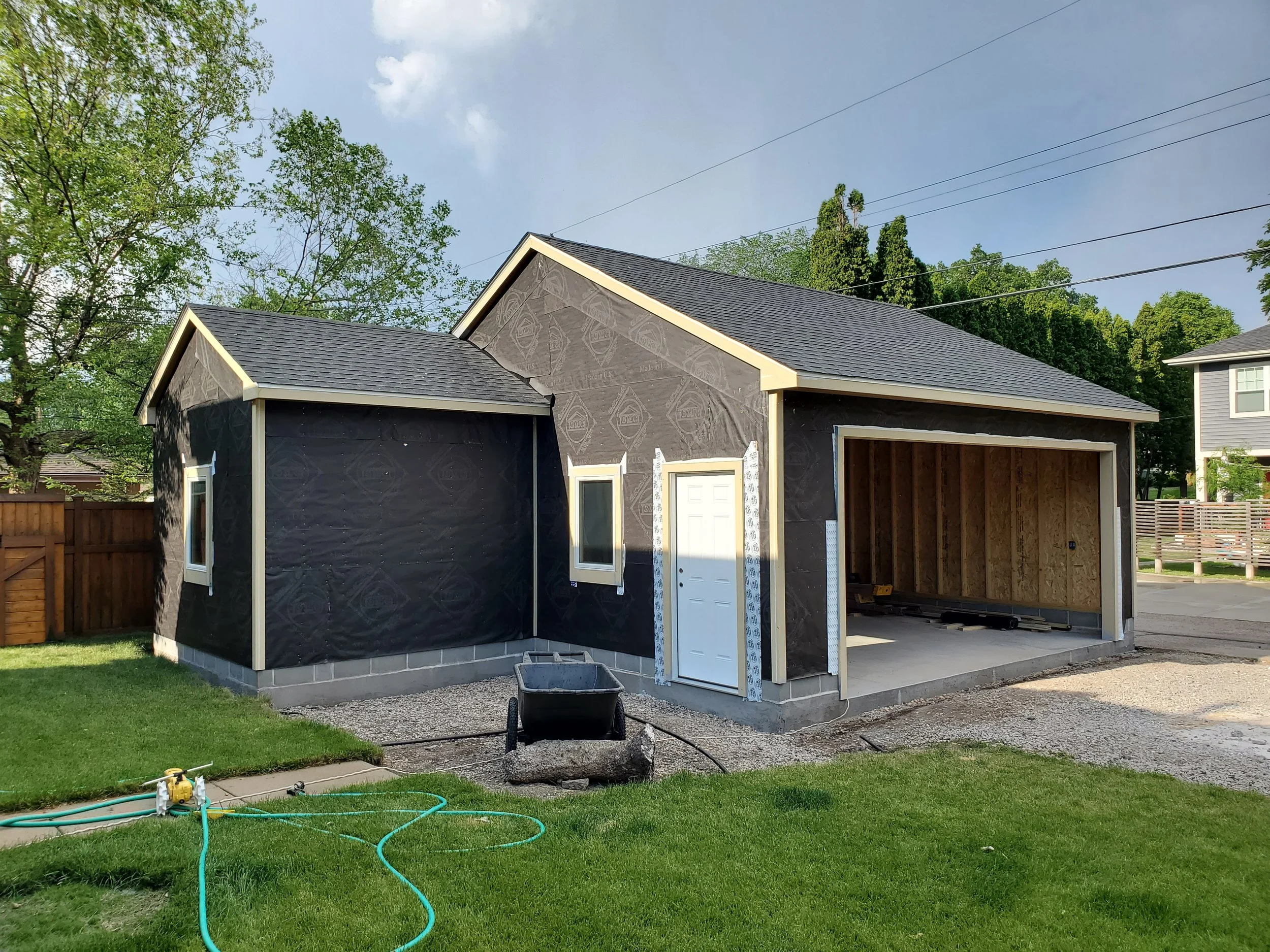 Newly constructed house with black roofing, black house wrap, and an unfinished garage, surrounded by a green lawn and wooden fencing.