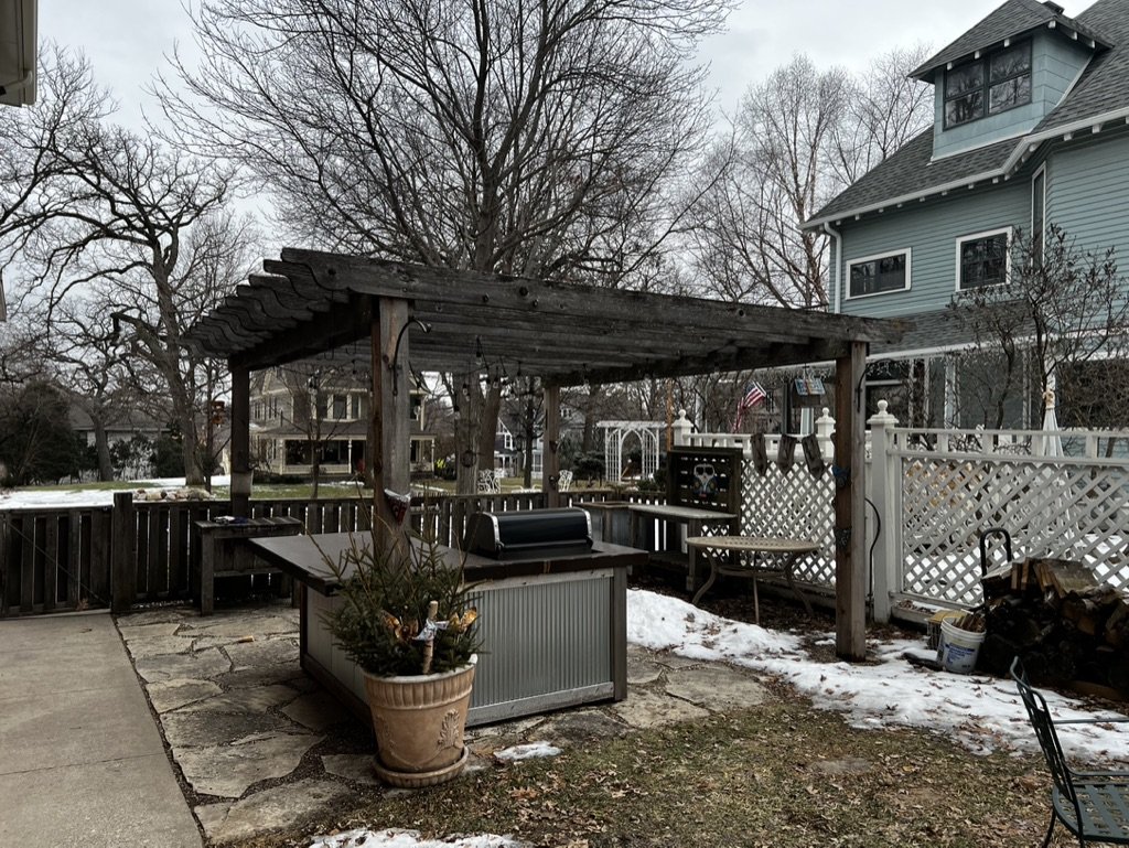 Backyard patio with a wooden pergola, a small grill, a flowerpot with a plant, and some snow patches, surrounded by a fence and neighboring houses.