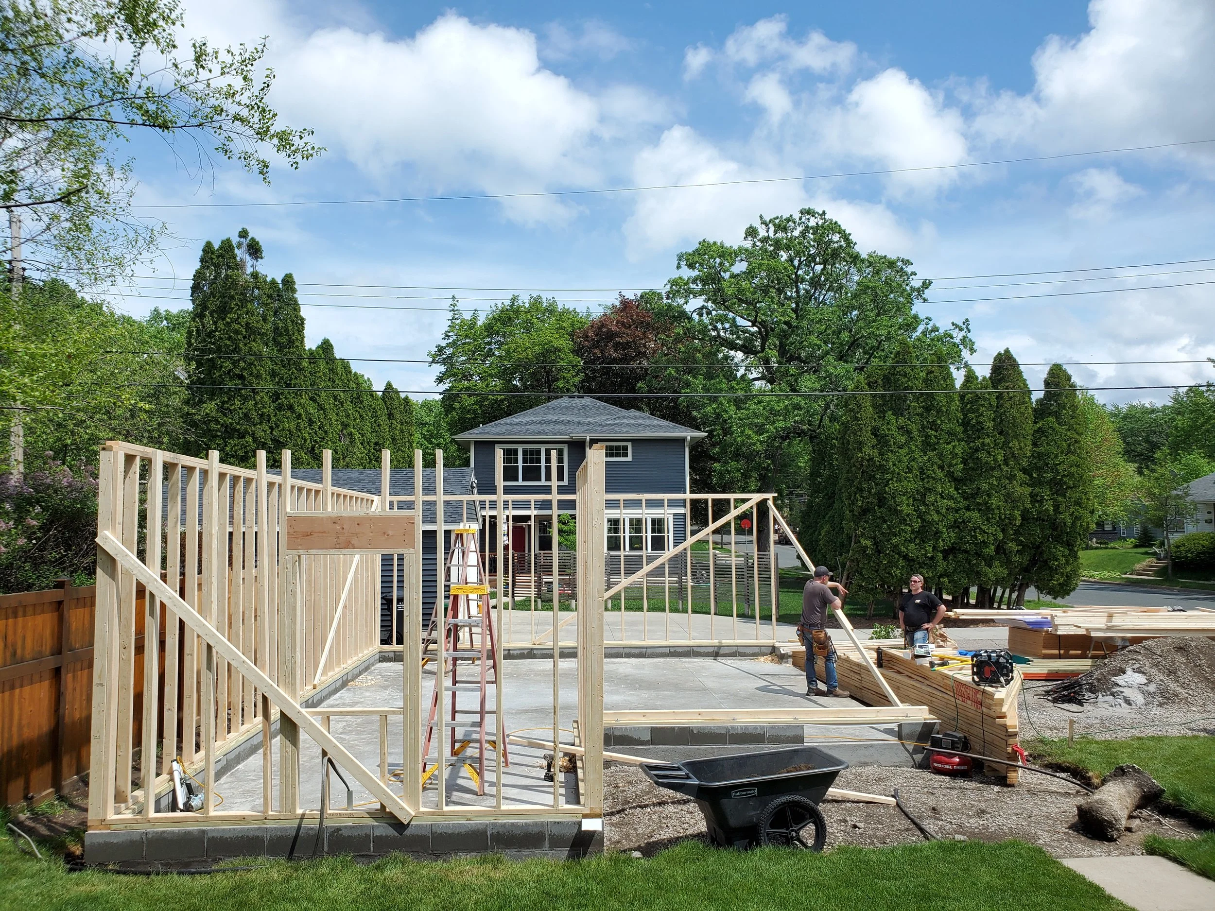 Construction workers building the wooden frame of a house in a suburban neighborhood with a blue sky and green trees in the background.