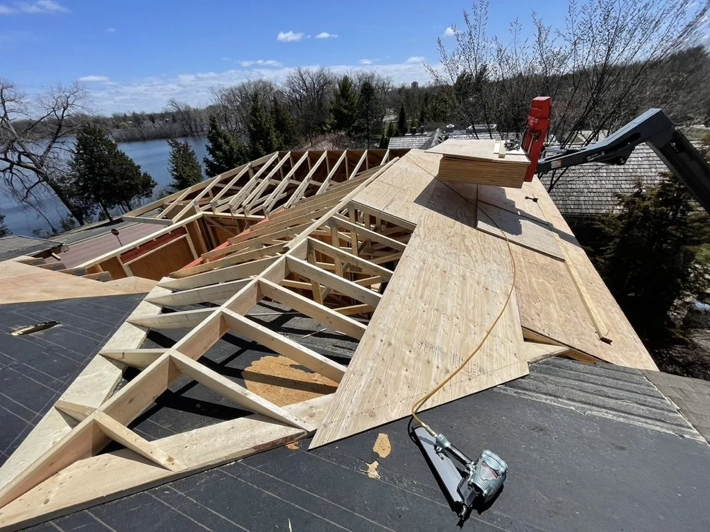 Construction site on a roof with wooden framing for a new roof structure, a nail gun, and stacked plywood sheets, with a backdrop of a lake, trees, and rooftops.