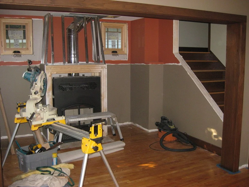 Interior of a room under construction with a saw, vacuum hose, and other tools, and a staircase leading to an upper level.