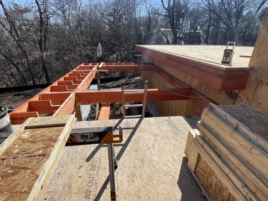 Construction site with wooden framing on a house, with a measurement tape on the roof, surrounded by trees.