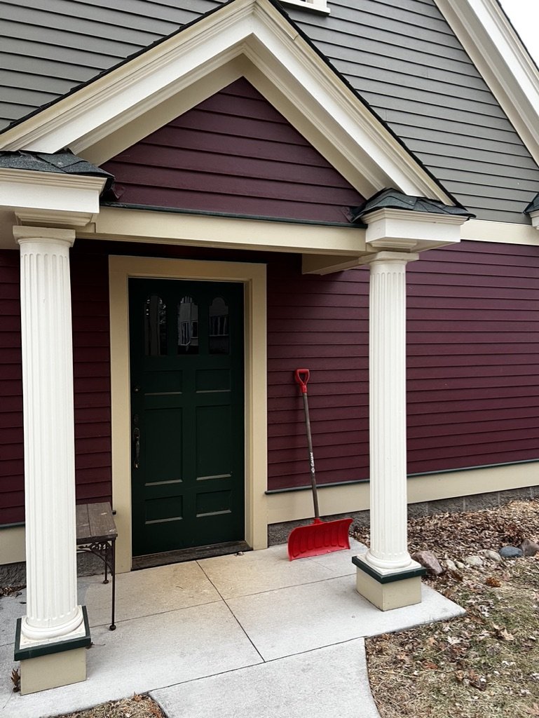 Front porch of a house with maroon and gray siding, white columns, a green door, and a red snow shovel leaning against the wall.