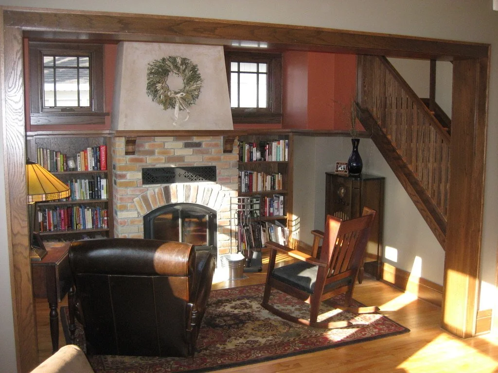 Cozy living room with brick fireplace, wooden bookshelves filled with books, a leather armchair, a wooden chair with a cushion, a small rug, and a wood staircase leading upstairs.