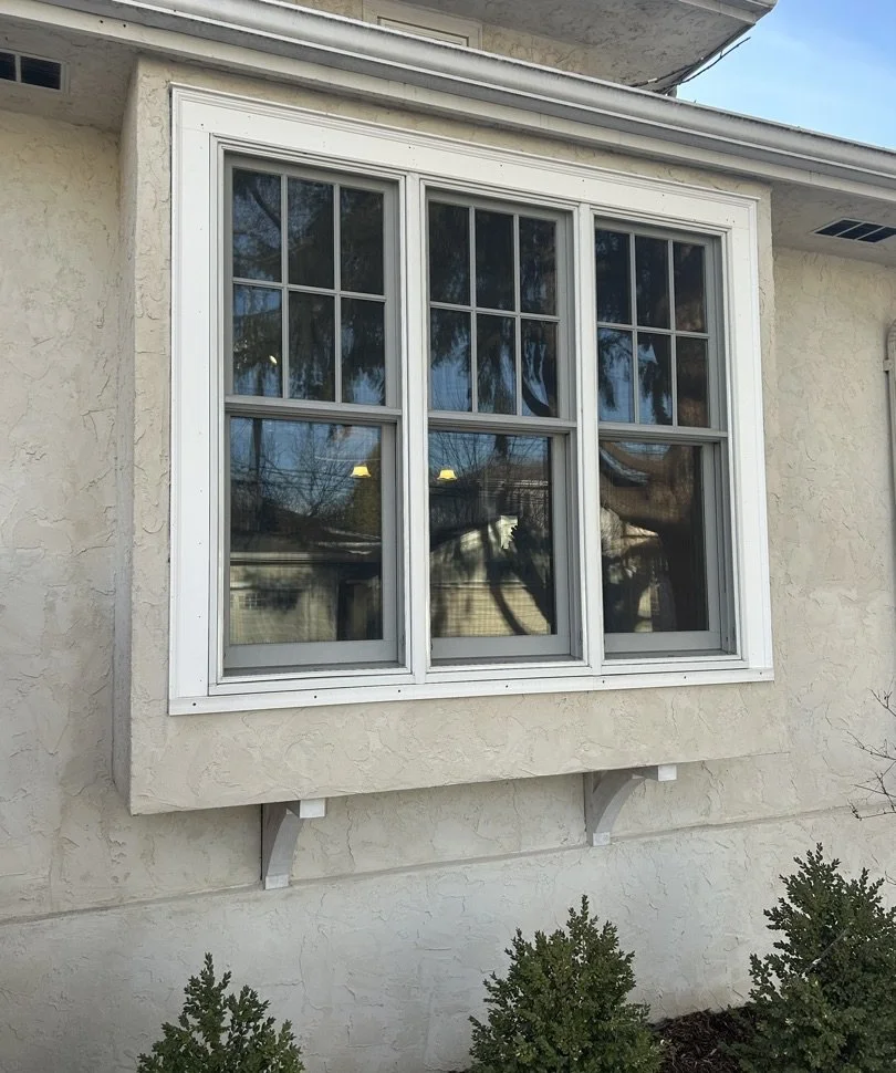 Exterior view of a house window with white framing, triple-pane glass, and decorative trim, set into a textured light-colored stucco wall.