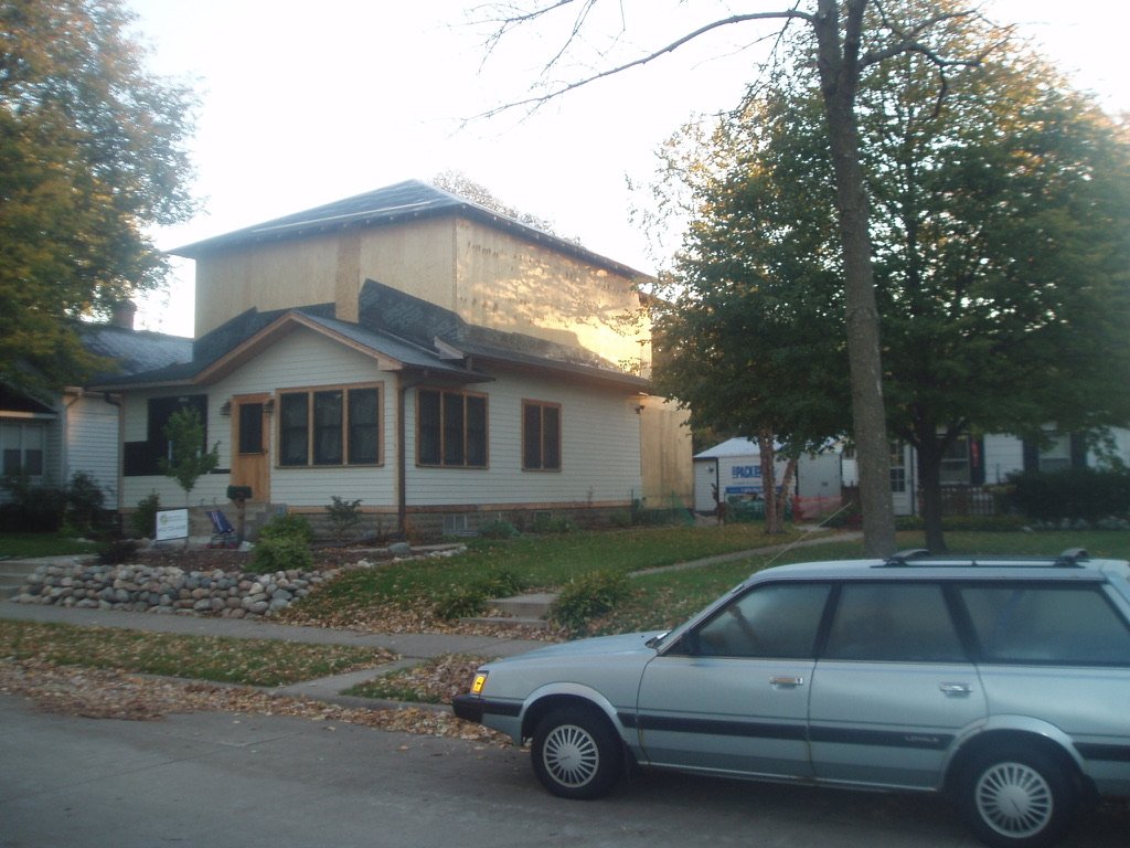 A house under renovation with an unfinished second story, surrounded by trees, with a silver station wagon parked in front.