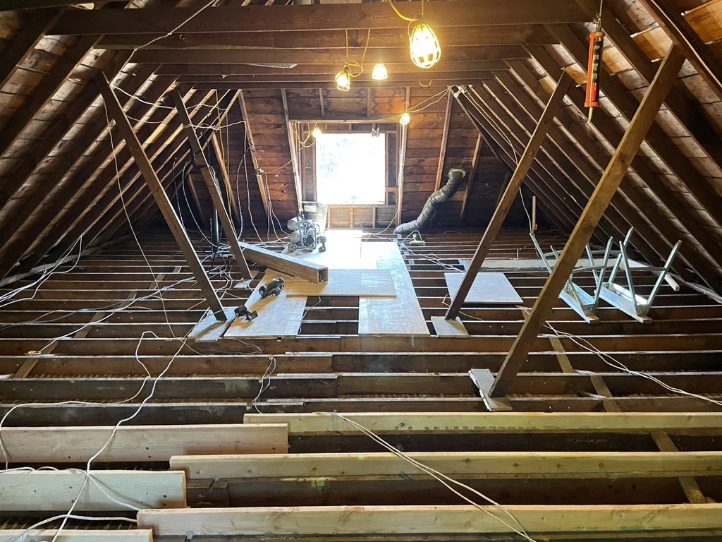 Attic space under renovation with wooden beams, electrical wires, a small window, and construction tools.
