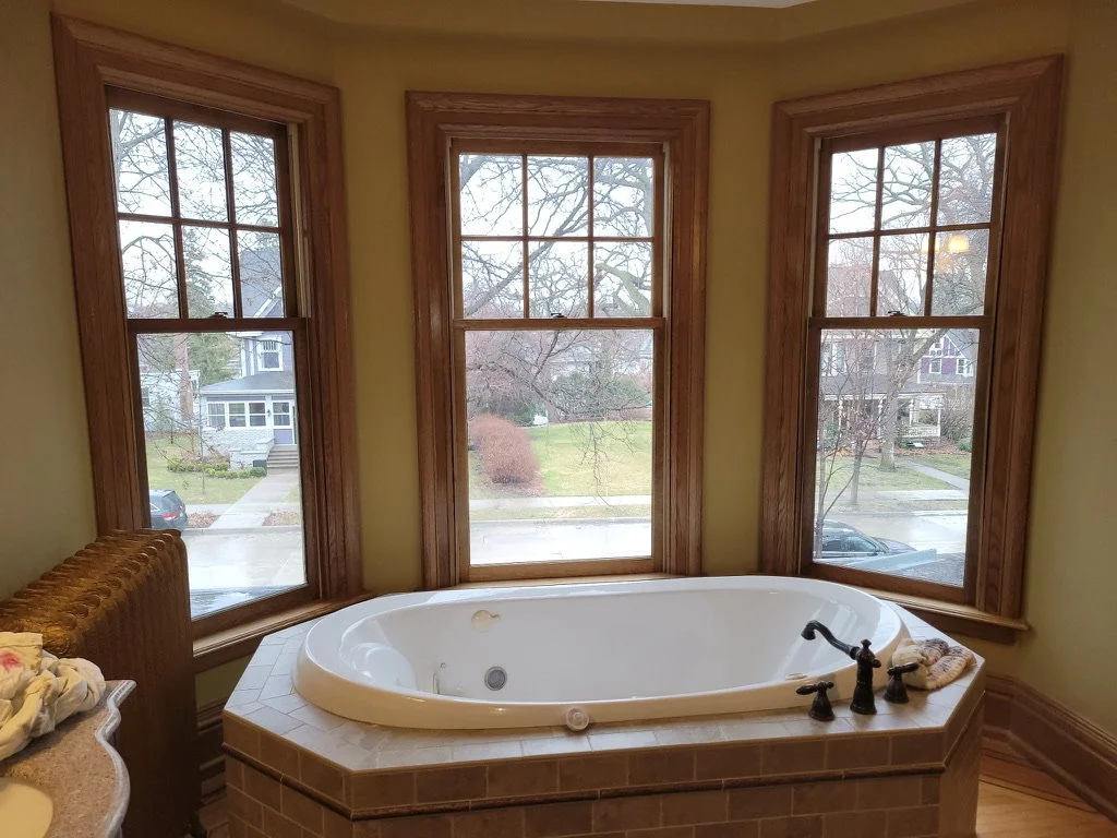 A bathroom with three large windows overlooking a suburban neighborhood, a white bathtub with black fixtures, and a small towel on the edge of the tub.