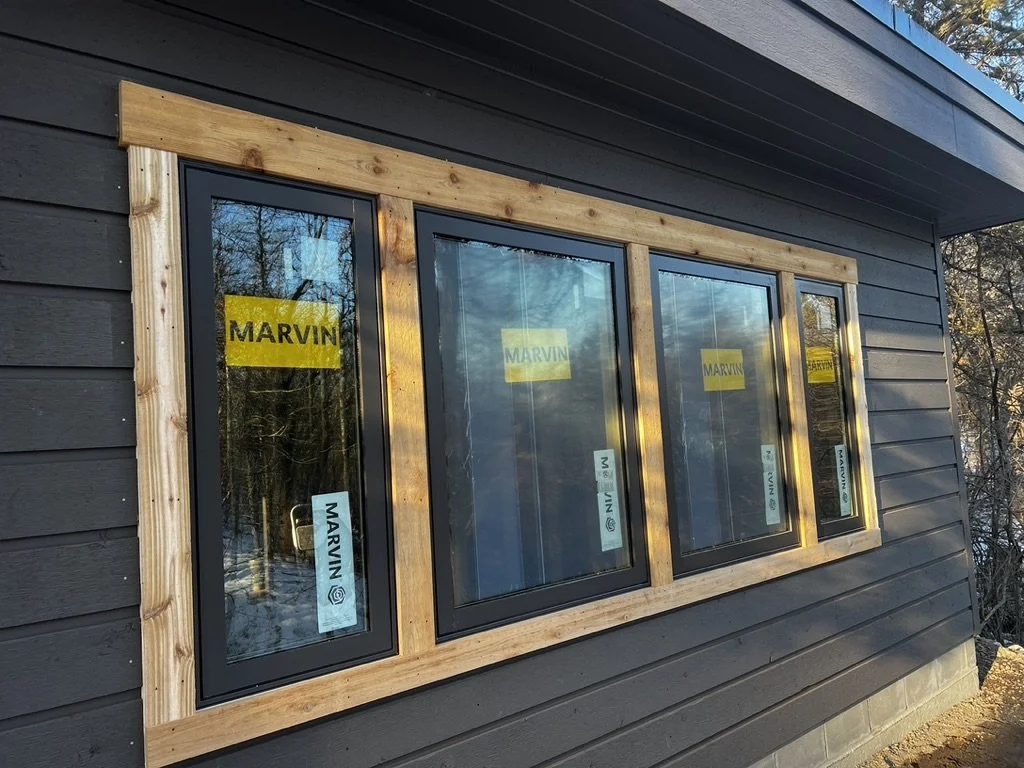 Four new double-pane windows installed on the exterior of a black wooden house. The windows are framed with unfinished wood, and yellow stickers with the brand name 'MARVIN' are visible on each window. The house has horizontal siding, and the backgro