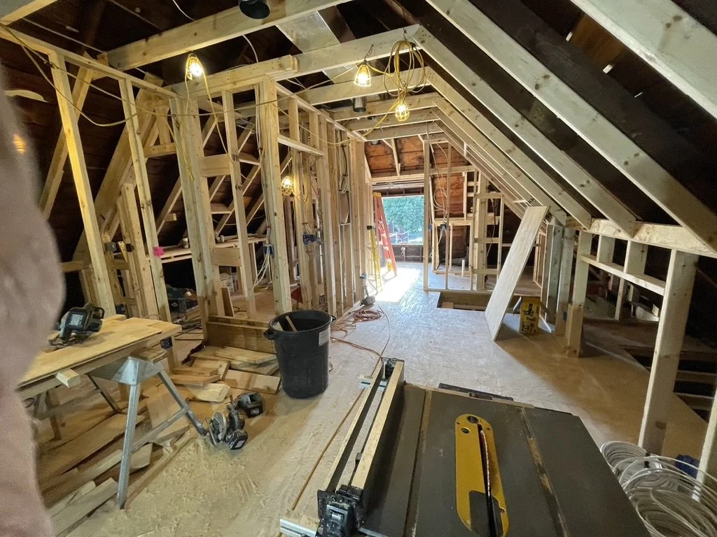 An attic under construction with exposed wood framing, hanging light bulbs, construction tools, and a workbench, leading to an open doorway at the far end.