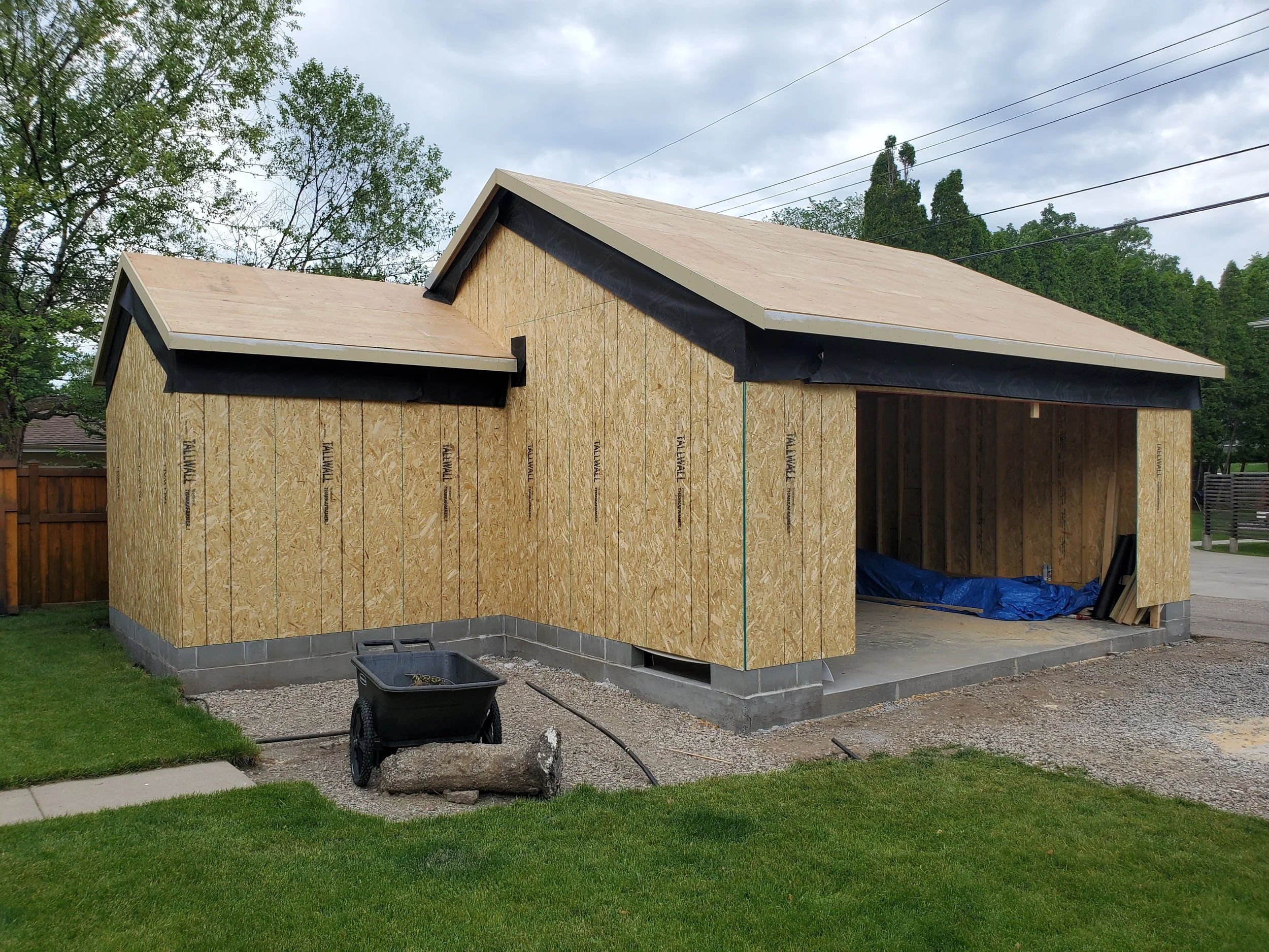 A house under construction with partially installed exterior walls made of plywood, and a roof with asphalt shingles. There is green grass, a black wheelbarrow, and construction materials around the site.