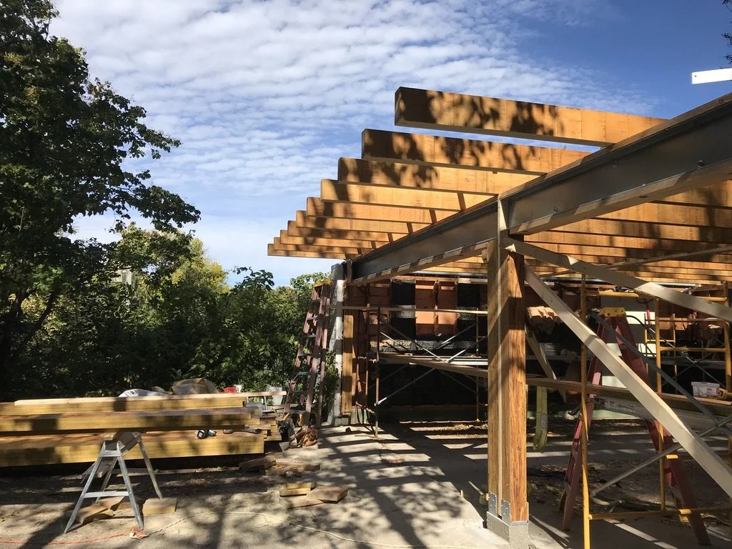 Construction site with wooden beams and scaffolding for a building extension, surrounded by trees and blue sky.